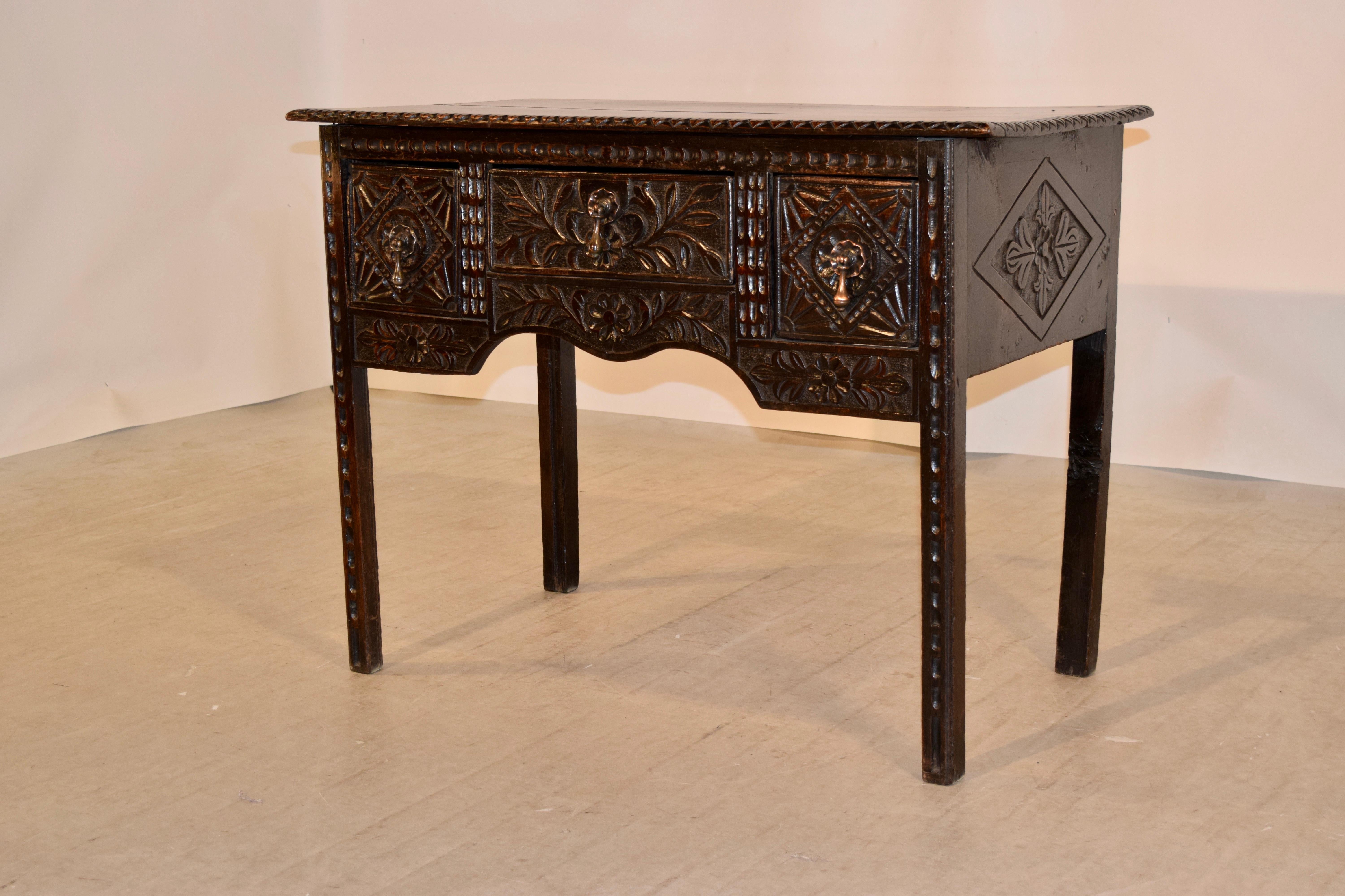 18th century oak lowboy from England with a two plank top which also has a beveled and carved decorated edge. This follows down to simple sides with carved medallion decorations and three drawers in the front, all with exquisitely hand carved
