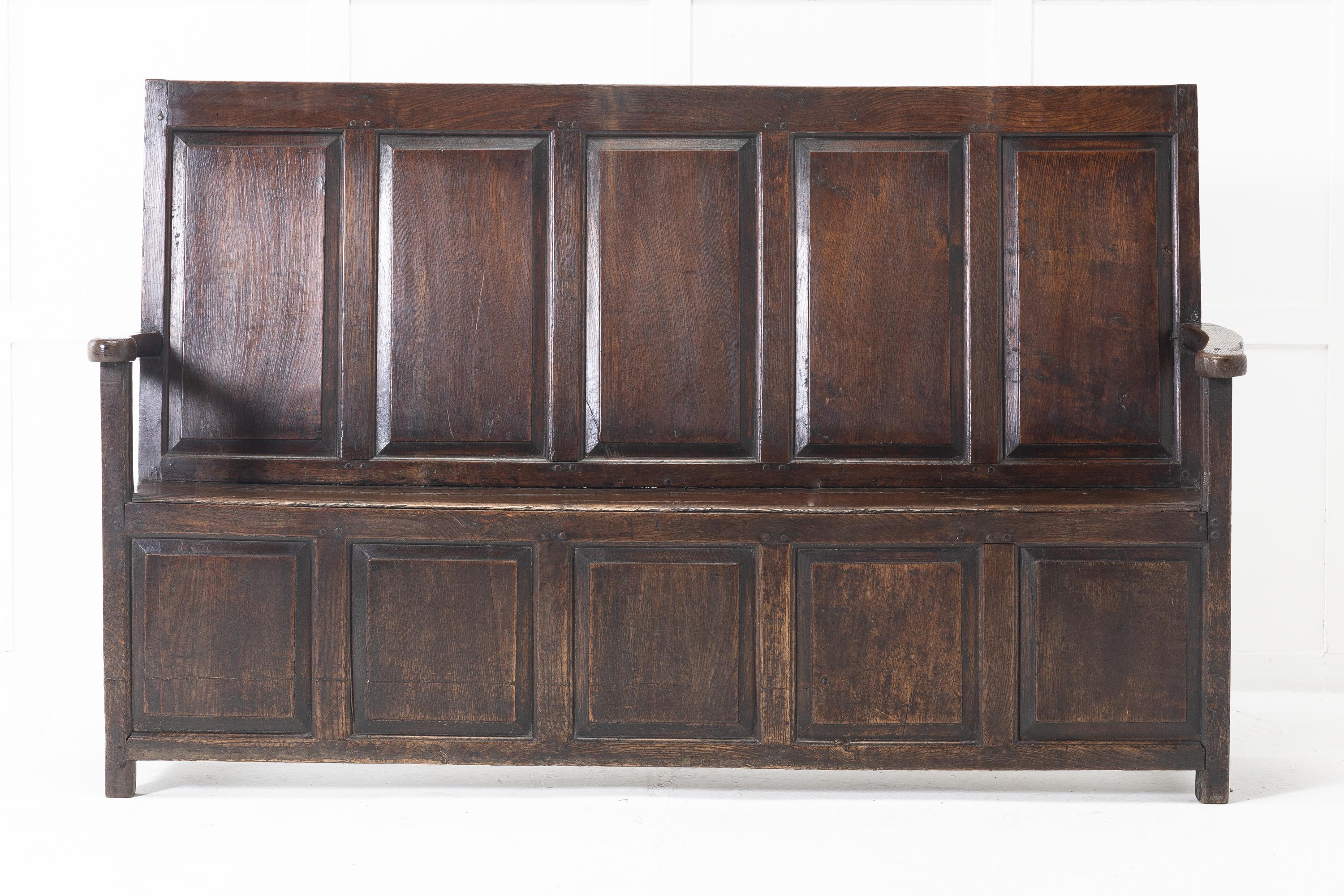 18th century English oak settle with a high back. Unusual format of five fielded panels at the back mirrored on the font panel, and having open arms.

Settles were generally placed by the large open fireplaces in country houses and inns. Usually