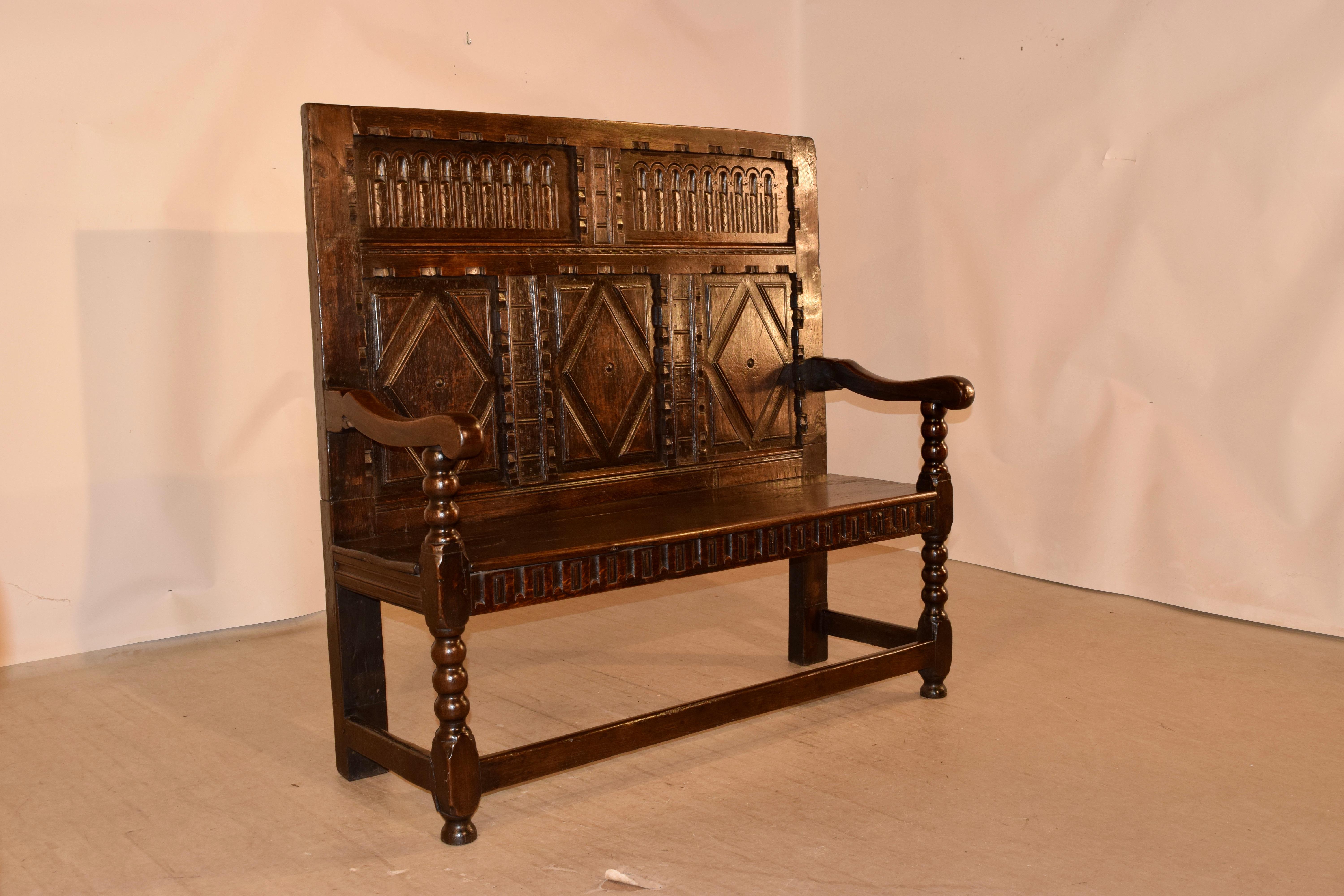 18th century oak settle from England with a wonderfully hand carved decorated and paneled back and rolled arms over hand turned bobbin arms and front legs. The seat height measures 18.75 inches and is over a hand carved apron. The legs are joined by