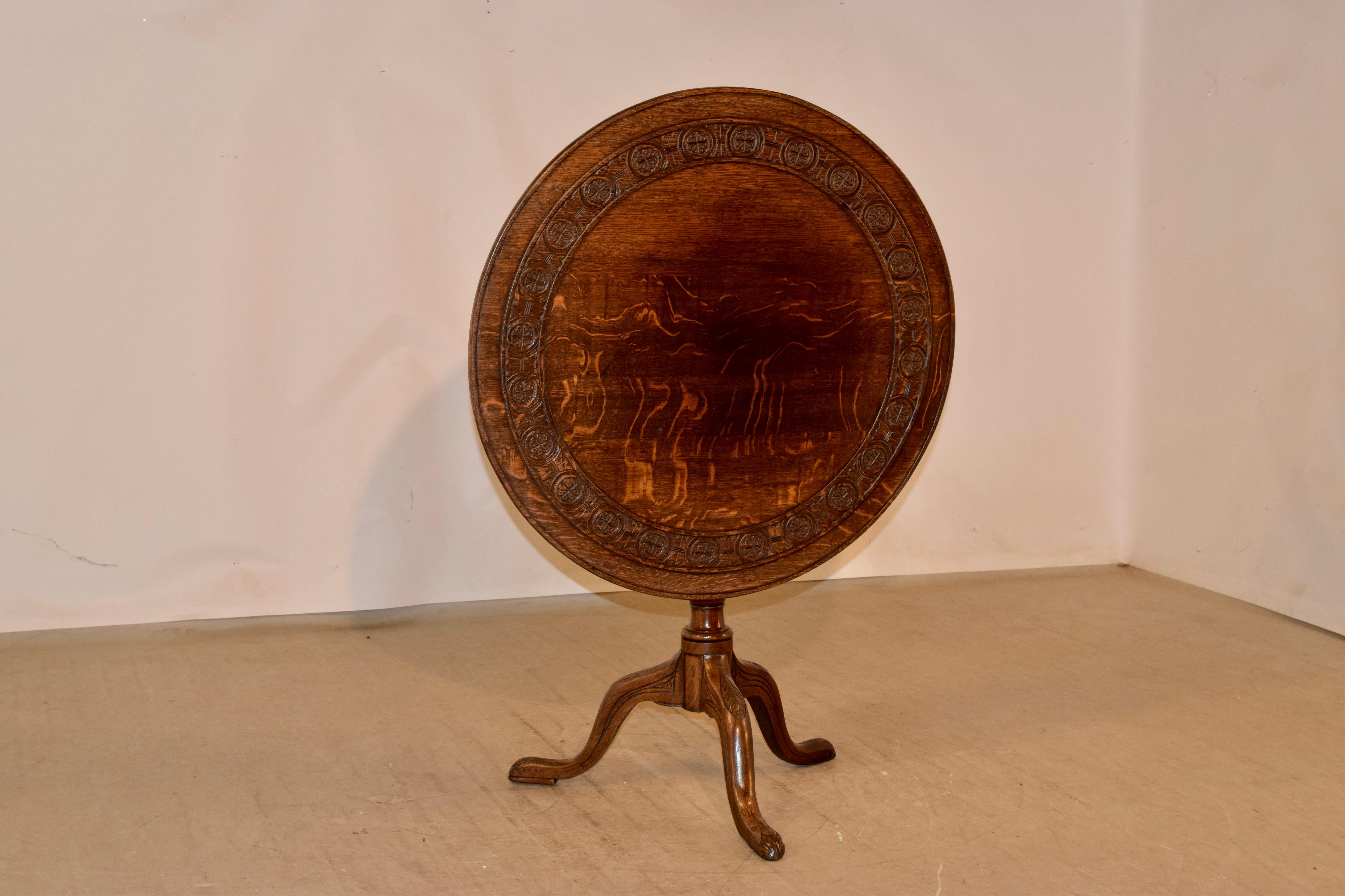 18th Century oak tilt-top table from England with a beveled edge, surrounding a hand carved band. The top rests on a hand turned vase shaped central column, supported on three legs, all with decorative hand carved accents. the feet are padded, and