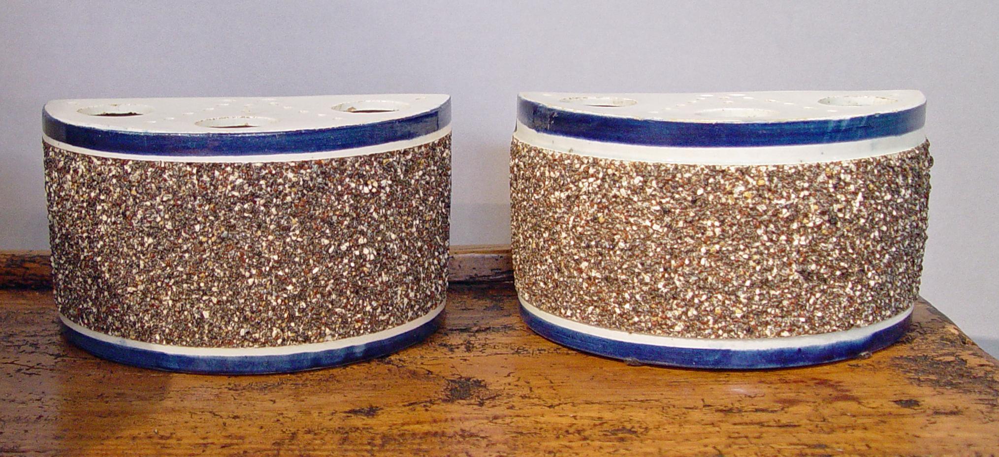 The pair of bough pots have a fixed pierced top with a blue band around the upper rim and the front panel with a pebbled or encrusted finish. The finish was created by applying small chips of crushed clay. These were designed to be displayed side by