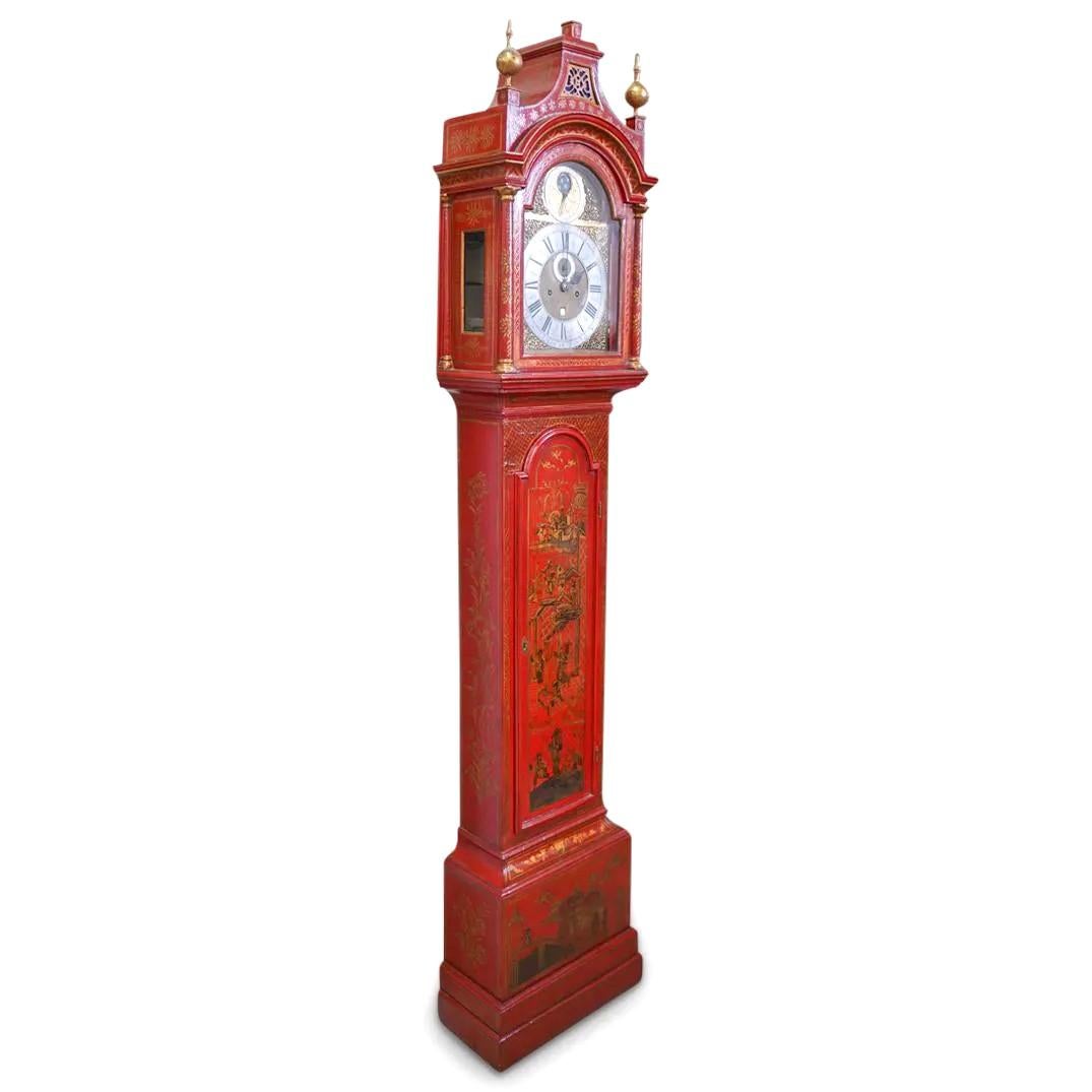 An 18th century English Chinoiserie grandfather clock or longcase clock. Features a red lacquered wood case with figural and village decorations. Clock housed in architectural arched case with gilt details and sphere finials. Clock face designed