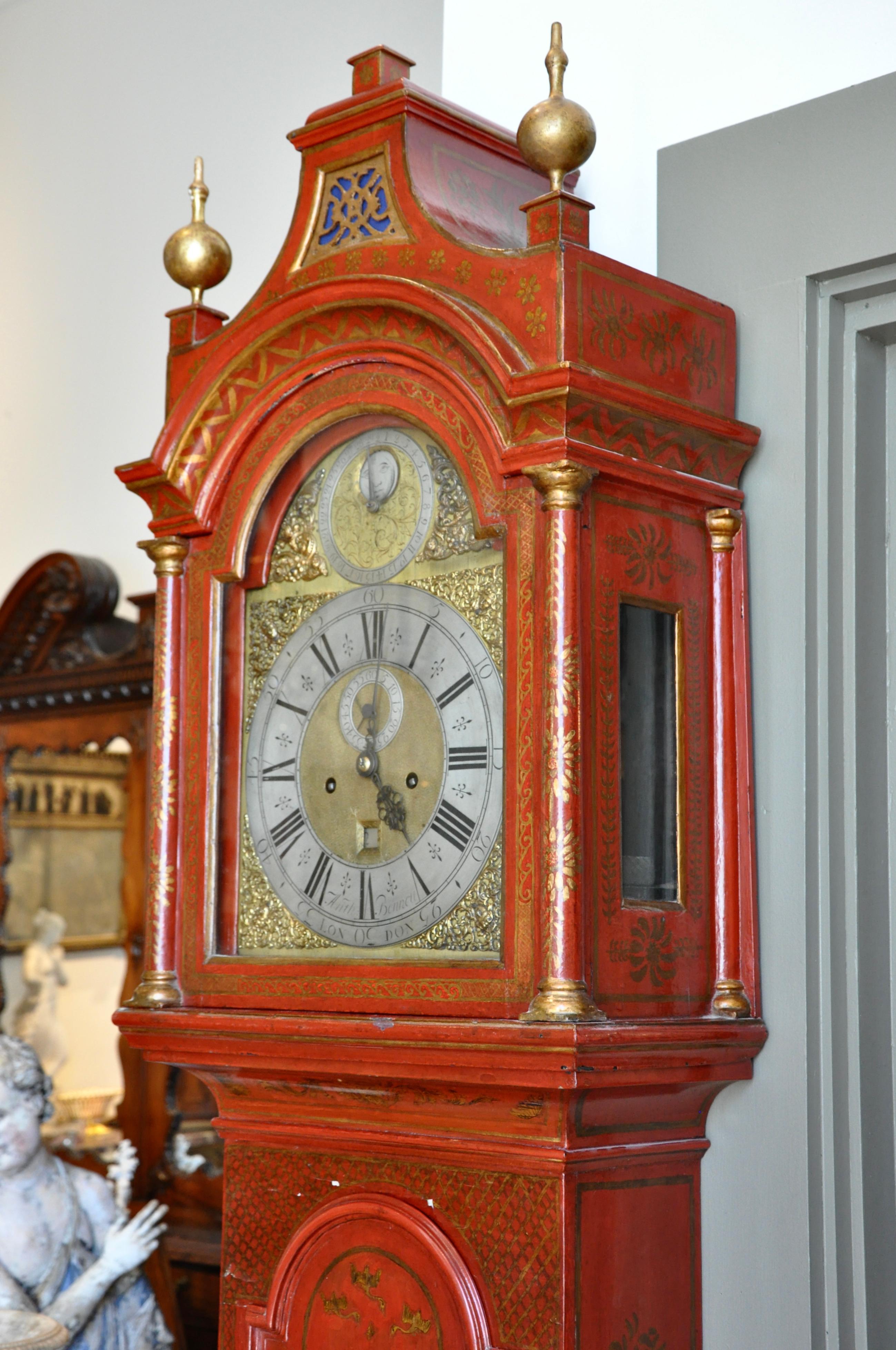 An 18th century English Chinoiserie grandfather clock or longcase clock. Features a scarlet red lacquered wood case with figural and village decorations. Clock housed in architectural arched case with gilt details and sphere finials. Clock face