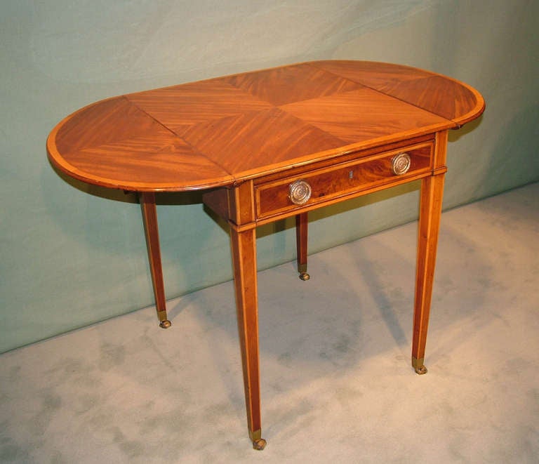 A late 18th century Sheraton period figured golden colored mahogany Pembroke table, satinwood crossbanded and boxwood and ebony strung throughout, having unusual quarter veneered 'D' ended top above frieze drawer supported on square tapering legs