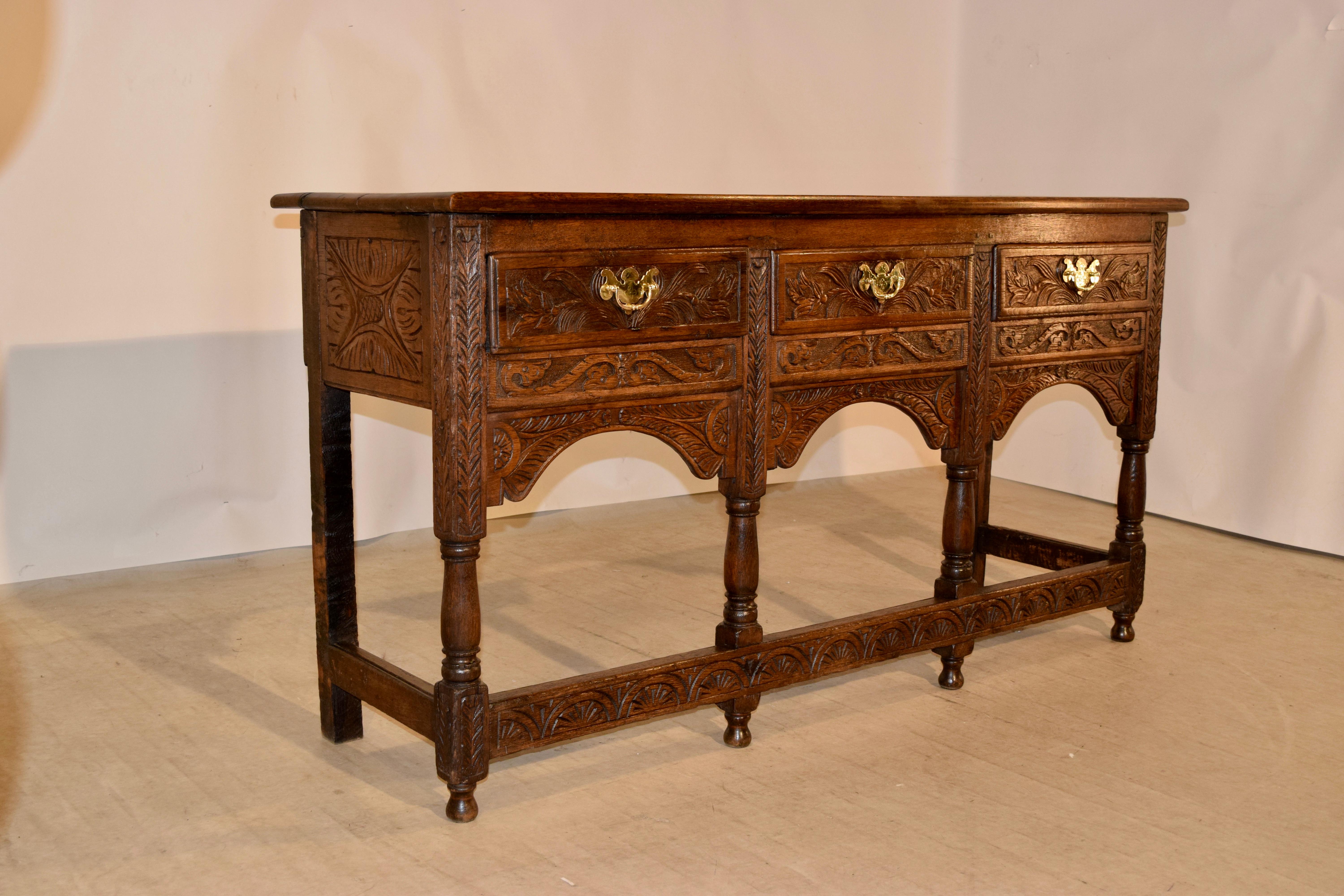 18th century English sideboard made from oak with three drawers, all with intricate hand-carved decoration and hand cast brass pulls with engraving over carved and scalloped aprons and carved panelled sides as well. The legs are hand turned and are