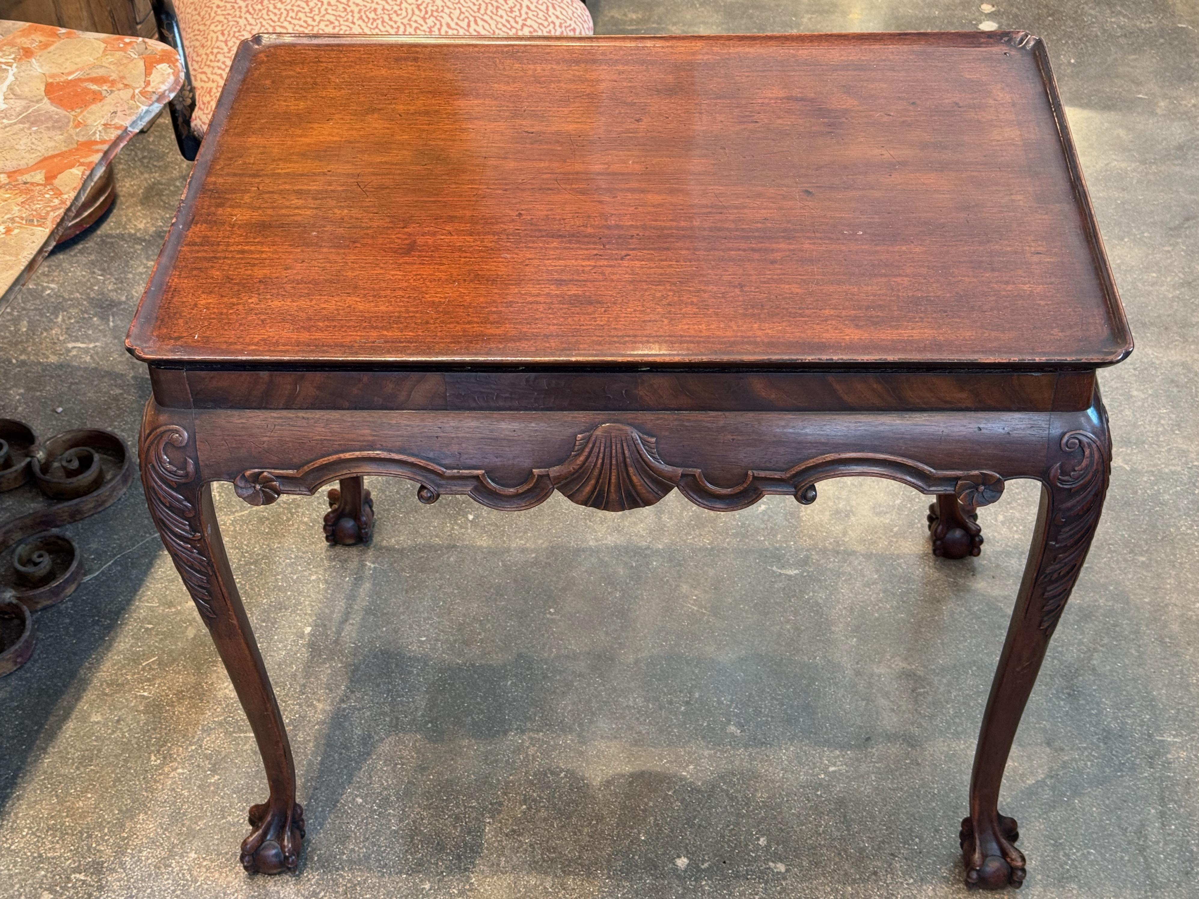 18th-century English tea table with a molded rectangular top featuring raised edges. The apron is adorned with a carved shell motif and scrolling details, complementing the cabriole legs with acanthus carvings and ball-and-claw feet. A refined and