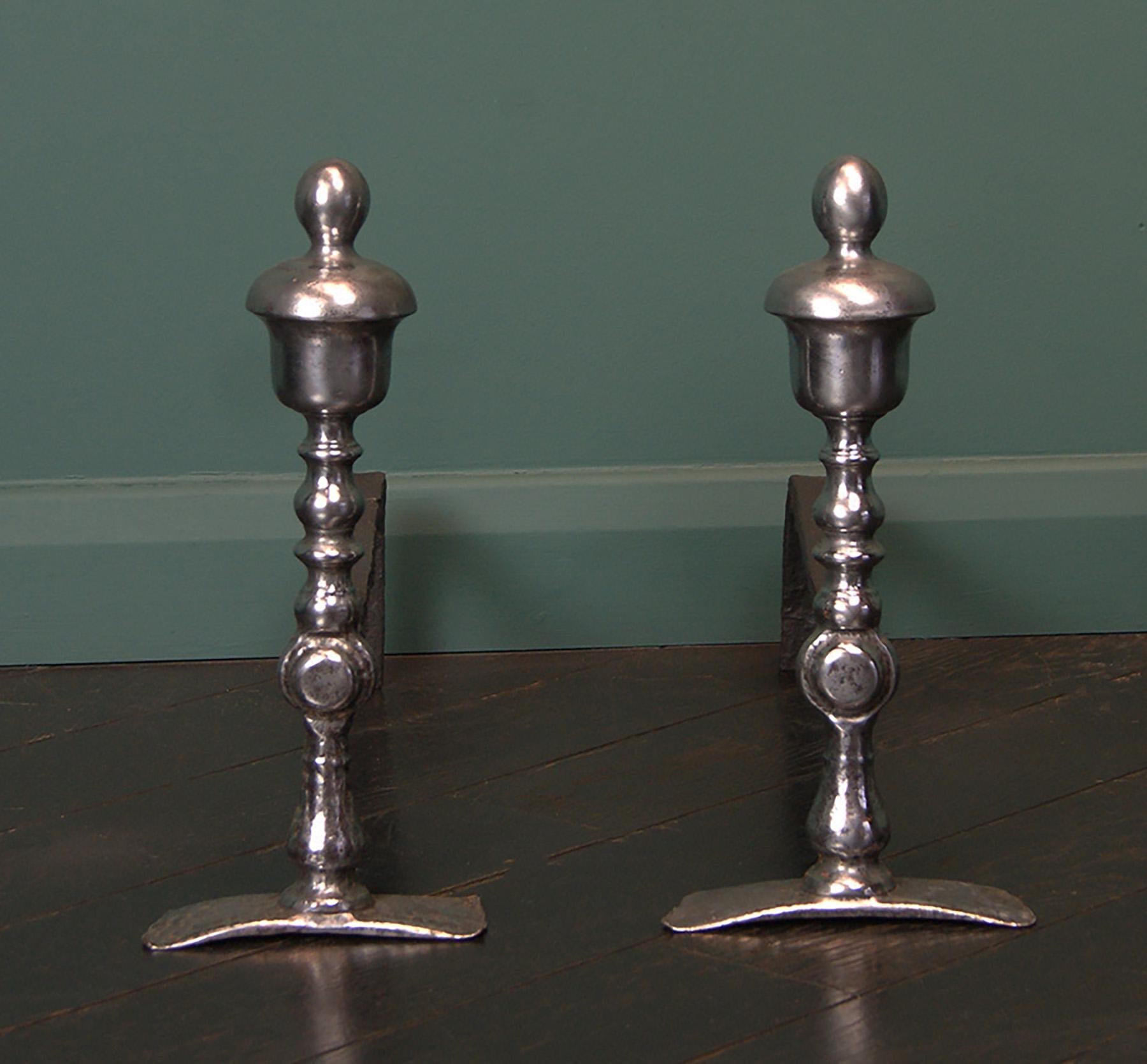 An early 18th century pair of English fireplace wrought-iron fire dogs. The wrought standards with heavy urn finials are riveted through with deeply veined paterna to rear log supports.  Restored.
Circa 1710