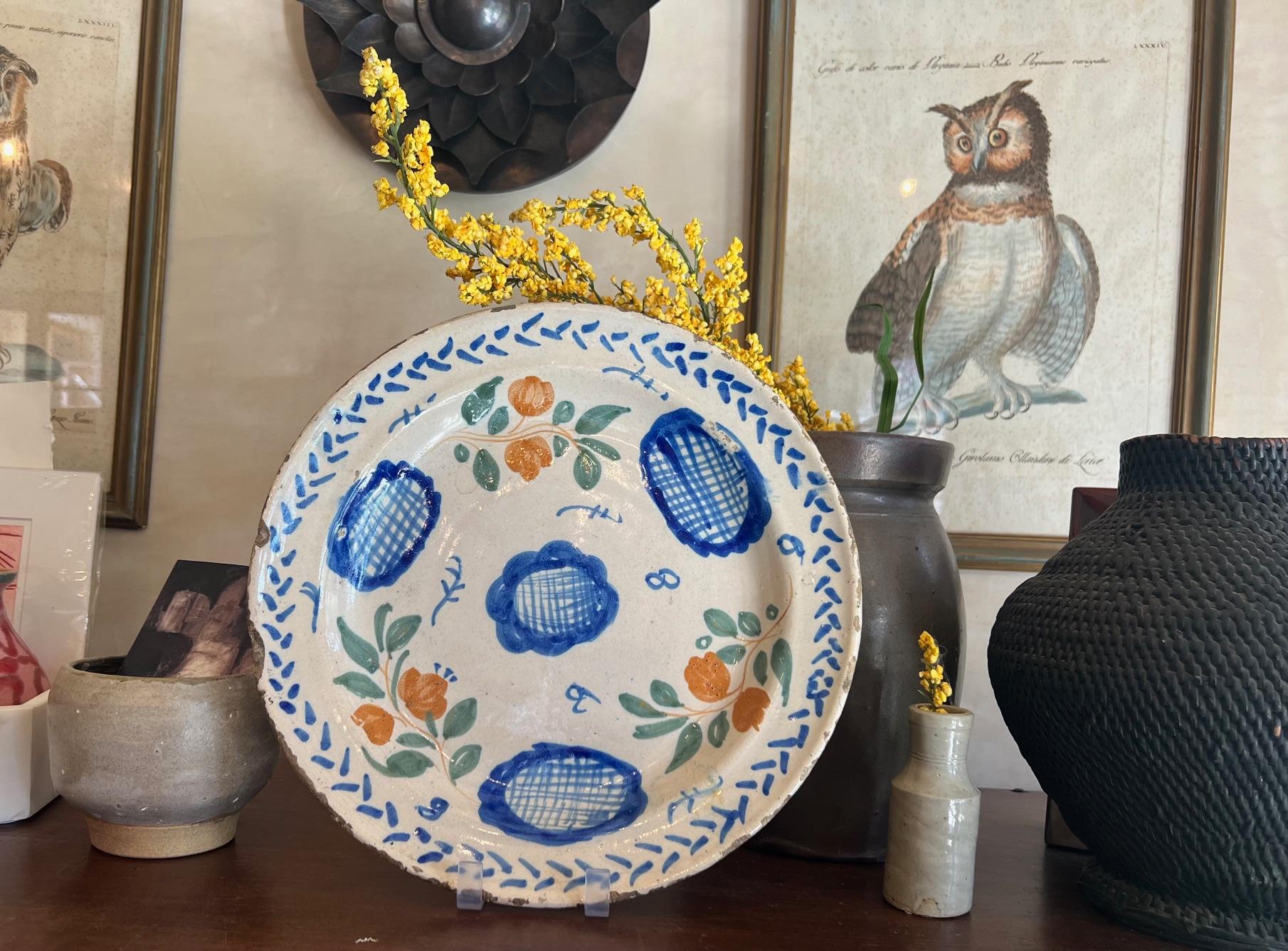 Simply painted European charger / plate / platter from the 18th century, likely Spanish. The tin-glazed, hand painted plate is decorated with blue and orange flowers surrounded by green leaves and blue accents.

I will include a plate hanger so it