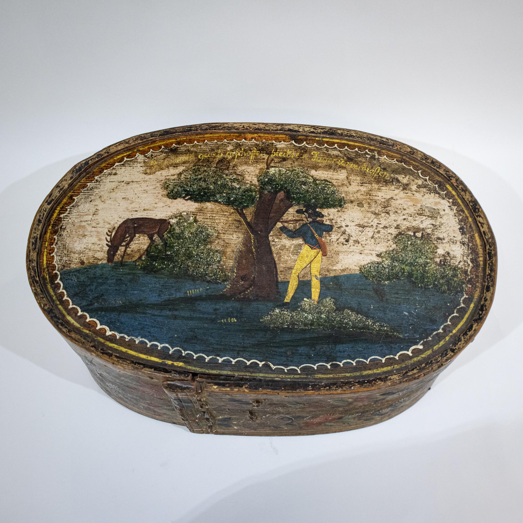 A lovely piece of Continental European folk art, this oval bentwood box has been painted with a landscape scene showing a huntsman with a rifle aiming at a stag or deer from behind a tree. There is some yellow text written in German around the top