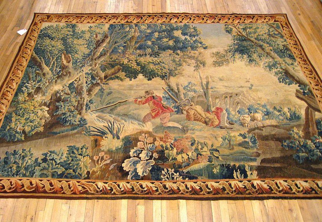 A Flemish pastoral hunting tapestry from the 18th century, depicting two noblemen seeking their catch between the large trees and verdant mounds of an idyllic forest setting, with a herd in the distance by the gateway to the village. Enclosed by a
