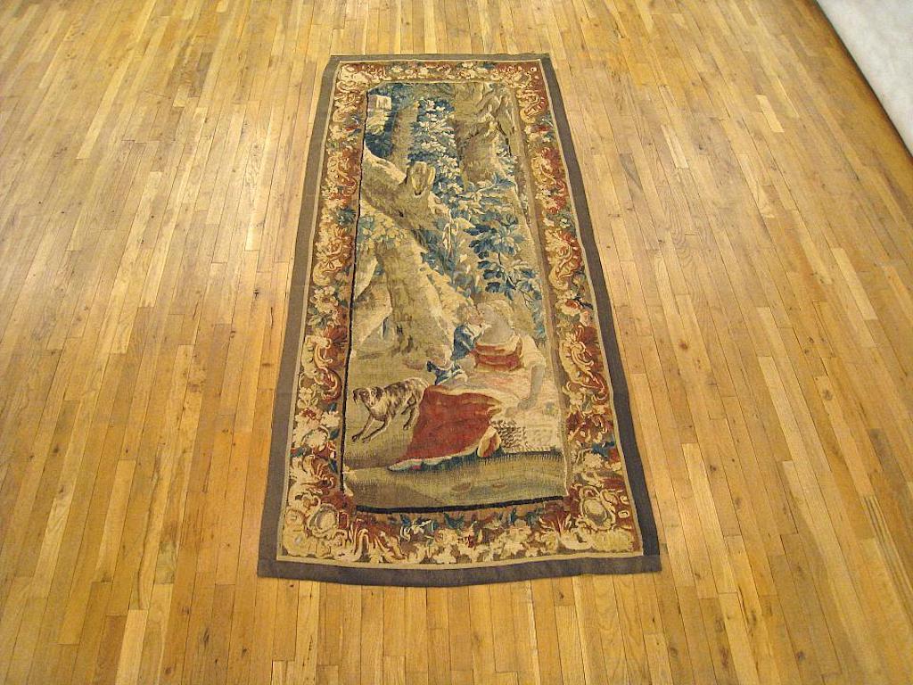 A Flemish rustic tapestry panel from the 18th century, featuring a lady reposing in shade of a large tree, seated with a picnic basket as her hound plays beside her. Enclosed by a scrolling floral border with intricate corner motifs. Wool with silk