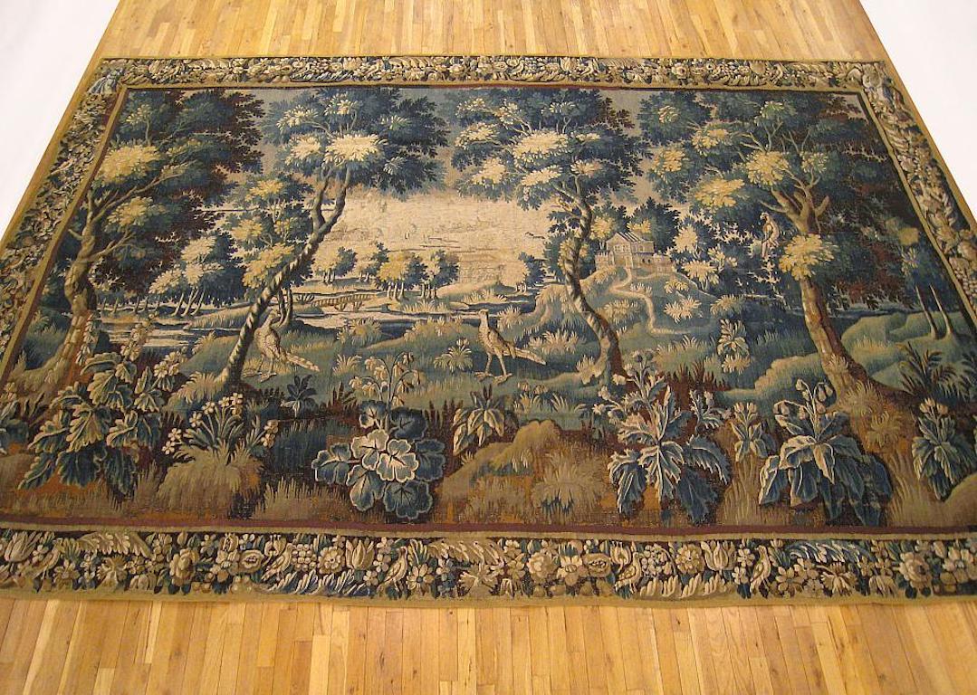 A Flemish verdure tapestry from the 18th century, depicting two exotic birds within a profuse landscape, with acanthus plants in the foreground, blooming bushes and trees at mid-ground, and a placid countryside along with elegant edifices in the