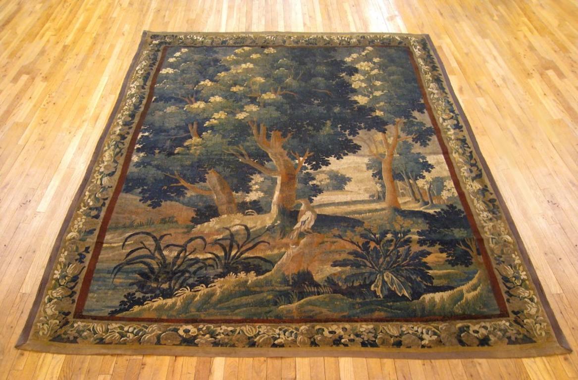 A Flemish verdure landscape tapestry from the 18th century, depicting a bird in a verdant scene between large trees and various foliage in front of a lake. Enclosed within a fruiting and flowering border. Wool with silk inlay. Measures: 9’10” H x
