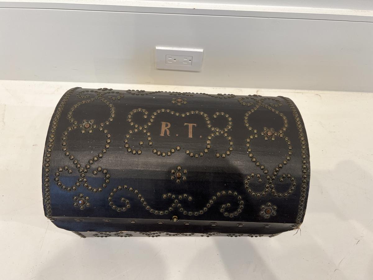 Se trata de una antigua caja de cuero del siglo XVIII, originaria de Francia. La caja está adornada con cabezas de clavo decorativas y lleva el monograma 