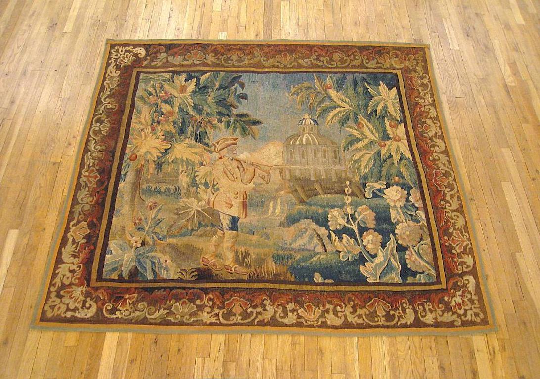 Panneau de tapisserie de genre d'Aubusson datant du XVIIIe siècle, représentant un chasseur marchant avec un fusil posé sur son épaule, avec un ruisseau et un château au loin, et la scène flanquée d'arbres en fleurs. Le tout est entouré d'une