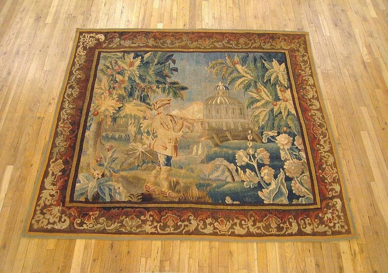 A French Aubusson genre tapestry panel from the 18th century, envisioning a hunter walking with a rifle resting on his shoulder, with a stream and a castle in the distance, and the scene flanked by flowering trees. Enclosed within a scrolling