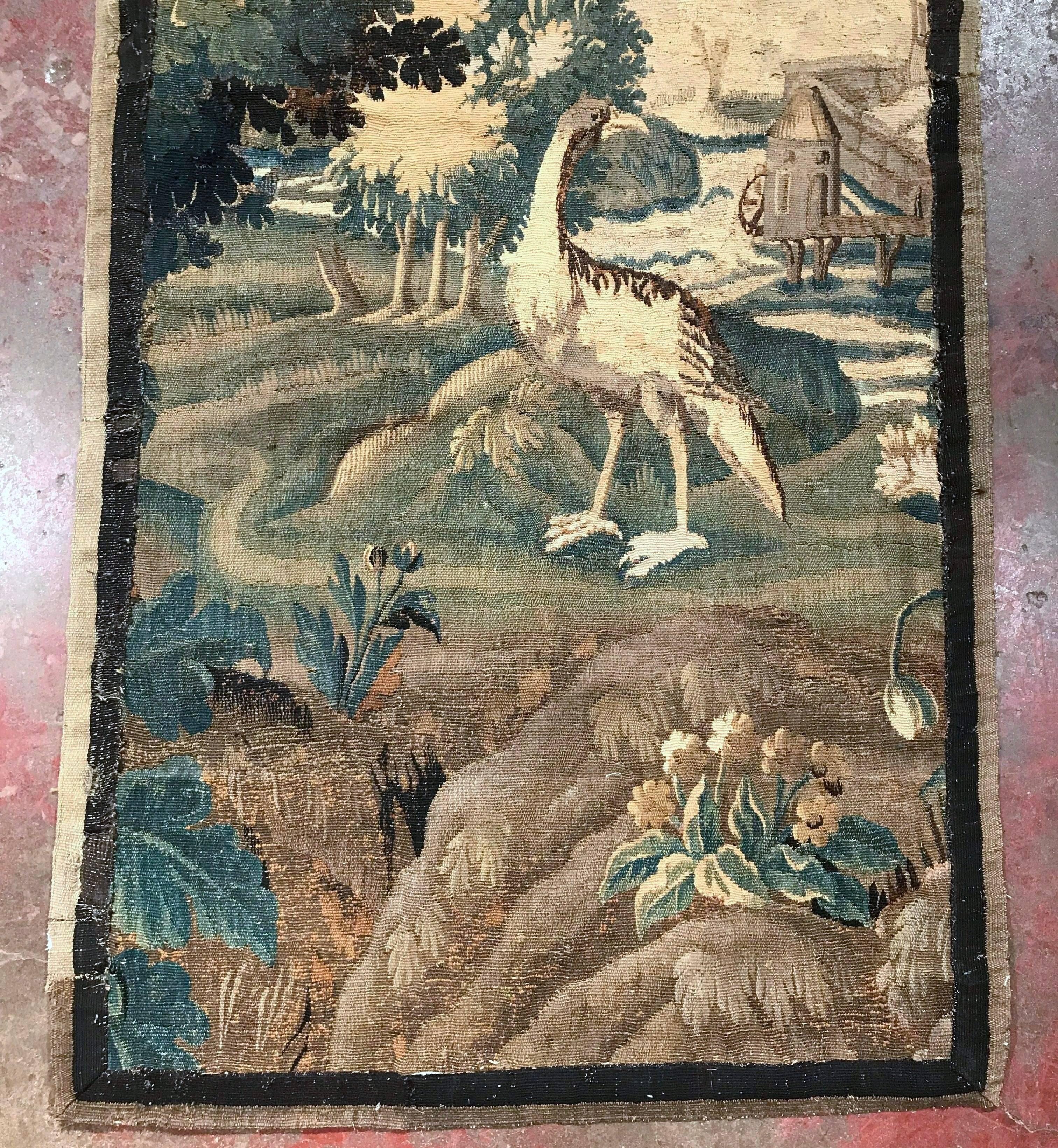 This tall and narrow antique tapestry fragment was handwoven in Aubusson, France, circa 1750. The colorful wall hanging verdure piece features a tall bustard (ostrich family) looking on, an exotic bird perched in a tree, and a windmill in the