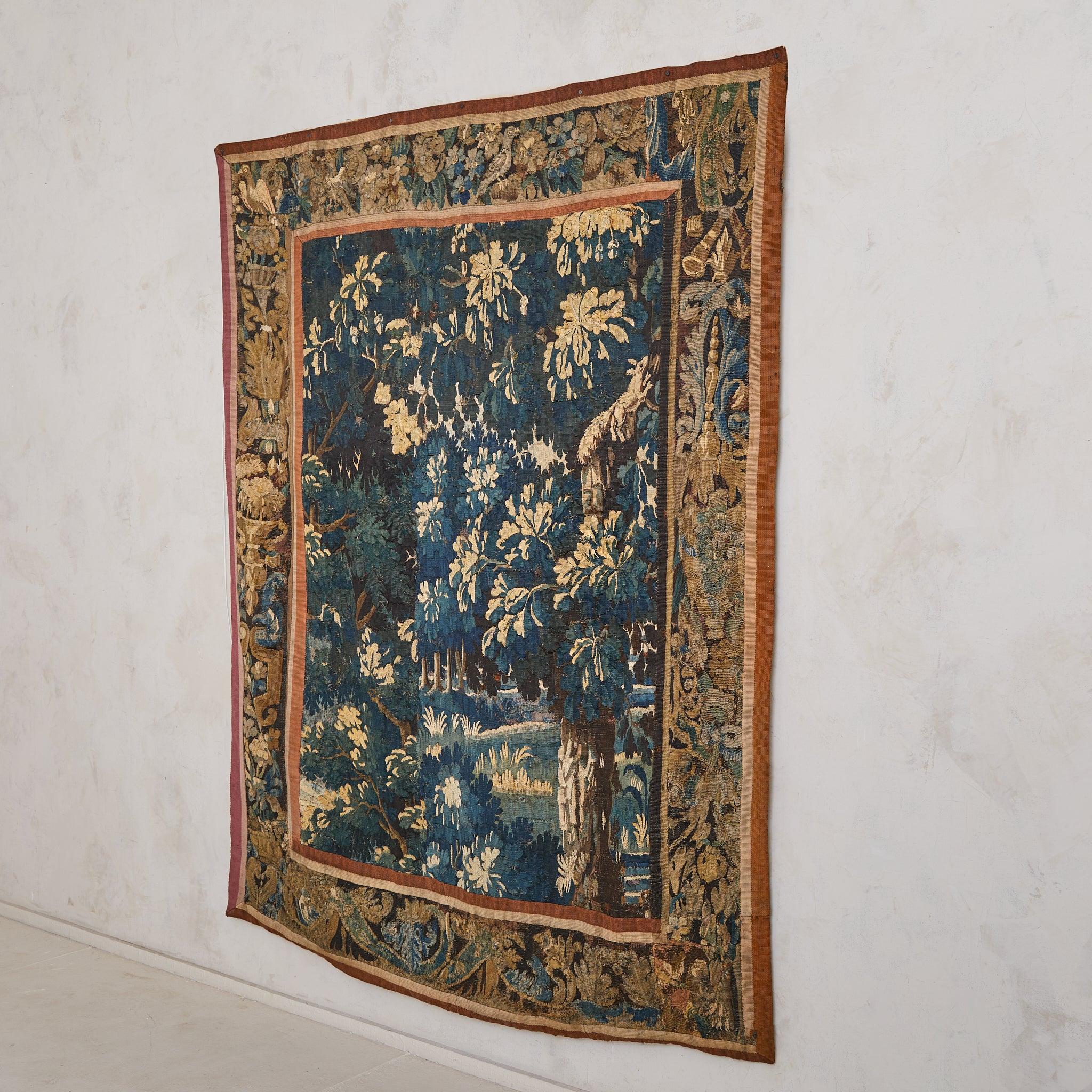 An 18th-century handwoven Aubusson tapestry depicting a verdant woodland in rich, moody tones. Dappled foliage cascades across the scene, rendered in deep indigos, mossy greens, and golden ochres—a pastoral dream suspended in time. A florid border