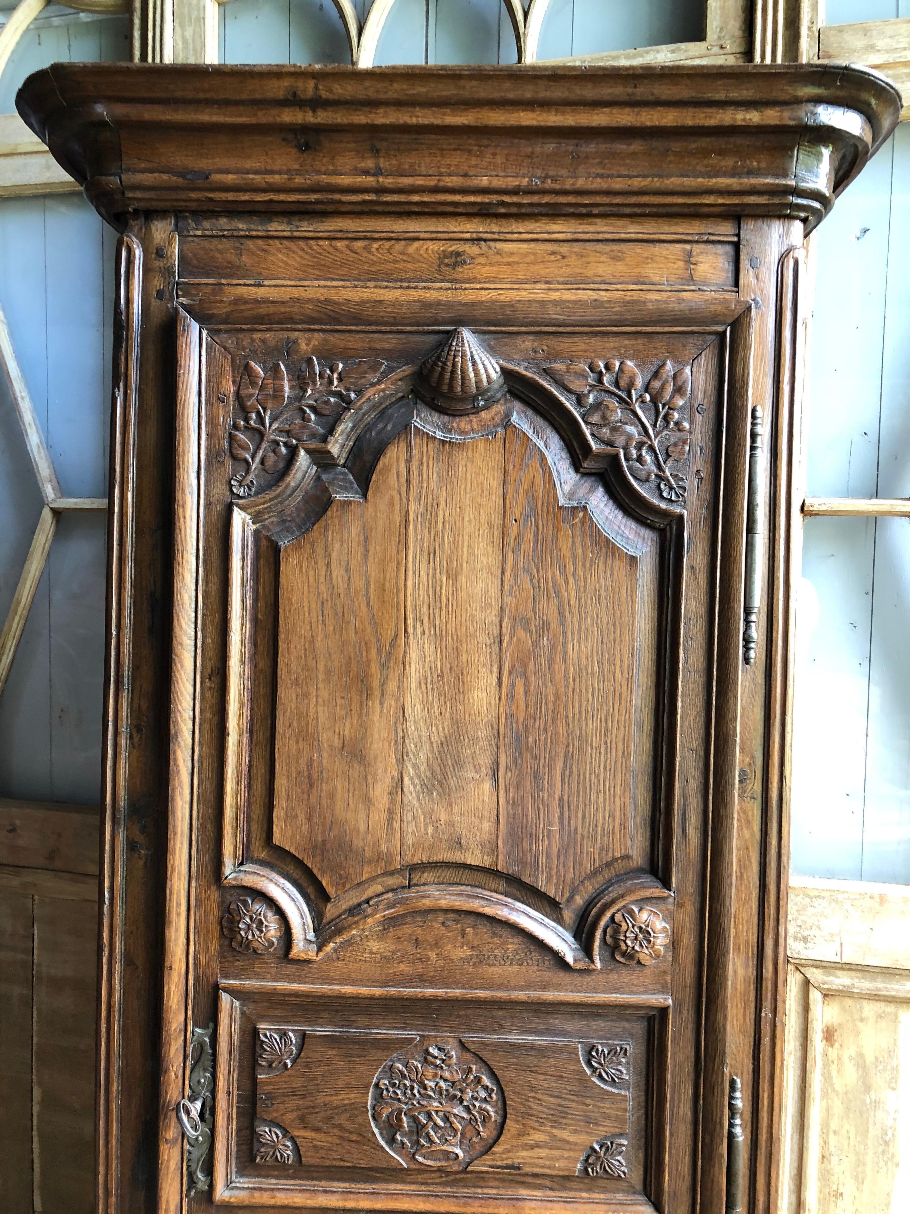 A mid to late 18th century French bonnetiere in oak, Normandy/Brittany region in the Louis XV style, with nicely carved decoration of flowers and a central scallop shell on the door, original lock and key and with 3 interior shelves. These narrow