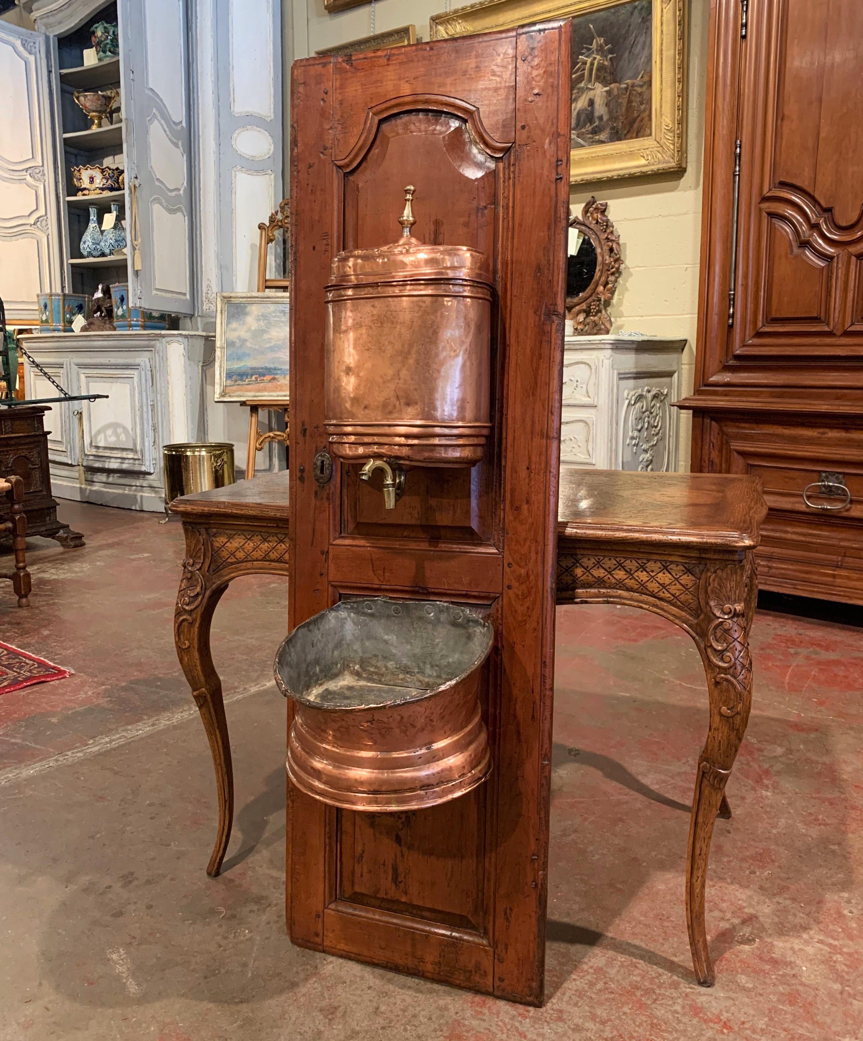Crafted in Normandy, France circa 1780, and mounted on a carved fruit wood door, the antique brass lavabo features the original two-piece lavabo; a demilune basin at the base, and a top reservoir with lid and brass faucet. The fountain is in