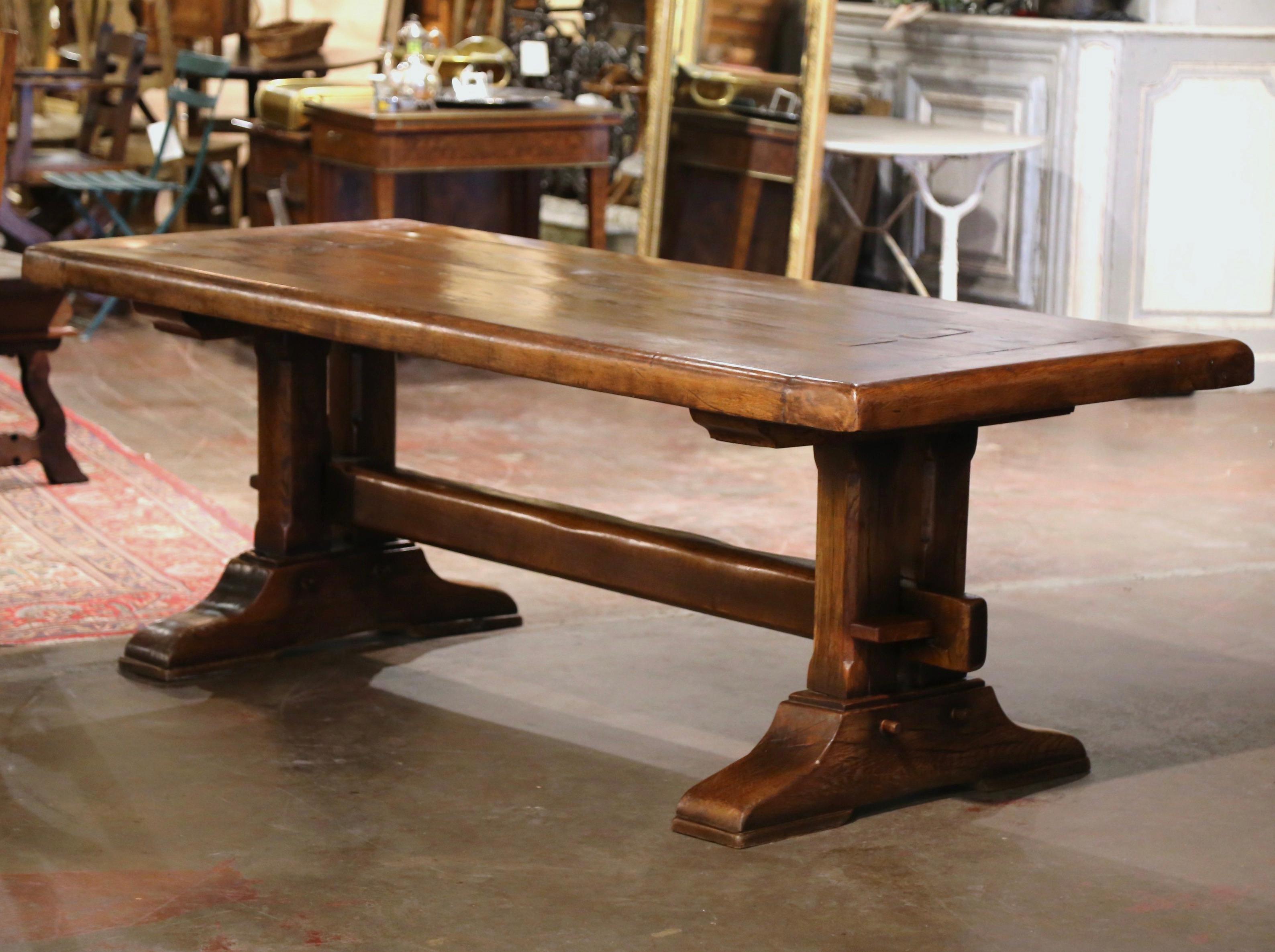 This elegant antique dining table was crafted in Normandy, France circa 1780. Built of solid oak, the refectory table stands on two carved trestle support legs ending with molded shoe base, and joined by cross stretcher attached with key and tenon.
