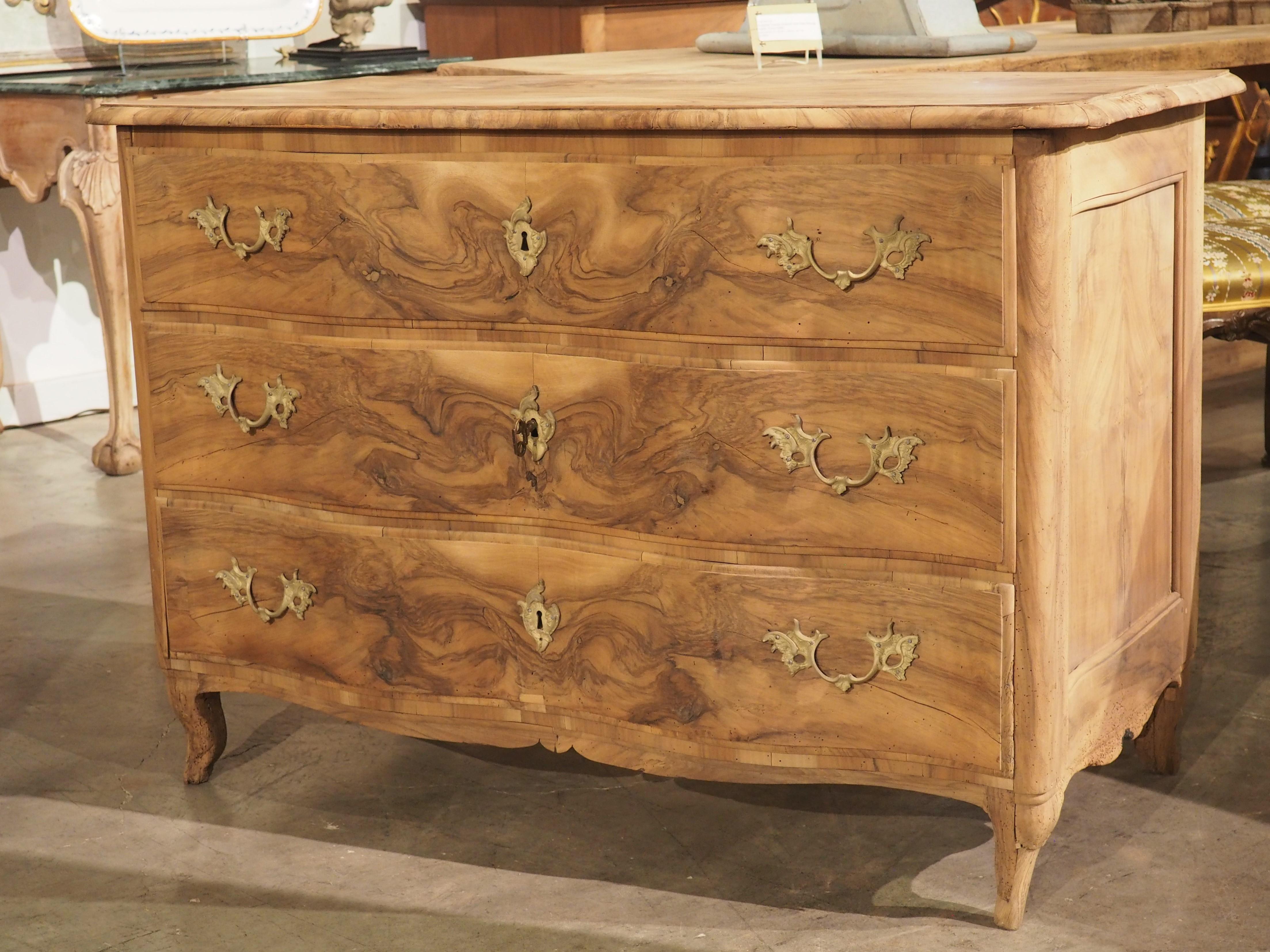 Hand-carved in France in the 1700’s, this arbalete front commode has been bleached at some point, revealing a fascinating burl walnut pattern. The chest of drawers has a warm, light brown color disrupted by the darker tones of the burl, which is