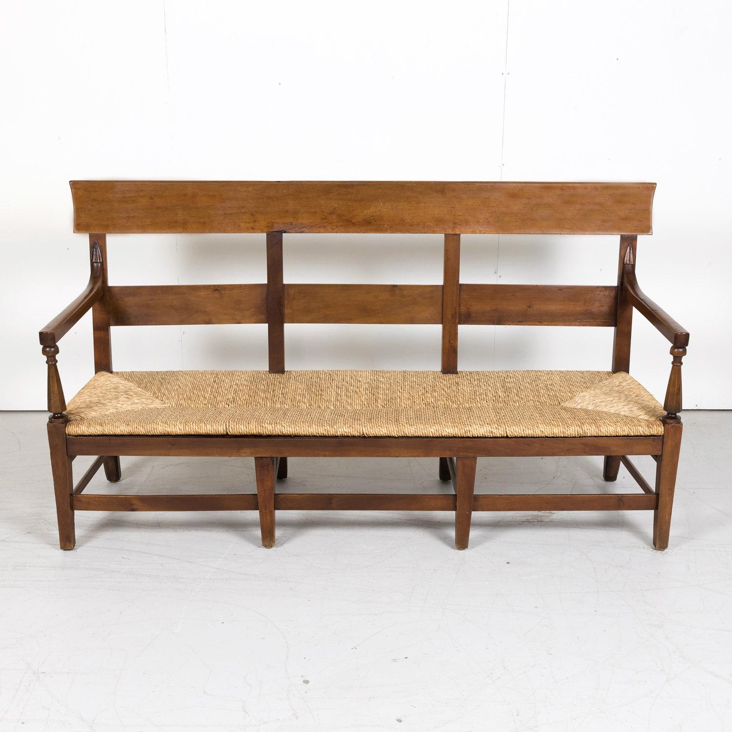 A handsome 18th century Country French Directoire period settee or radassier handcrafted in solid cherry near Marseille in the South of France, circa 1790s. This double arm bench seats three and features a handwoven rush seat, ladder-back, and