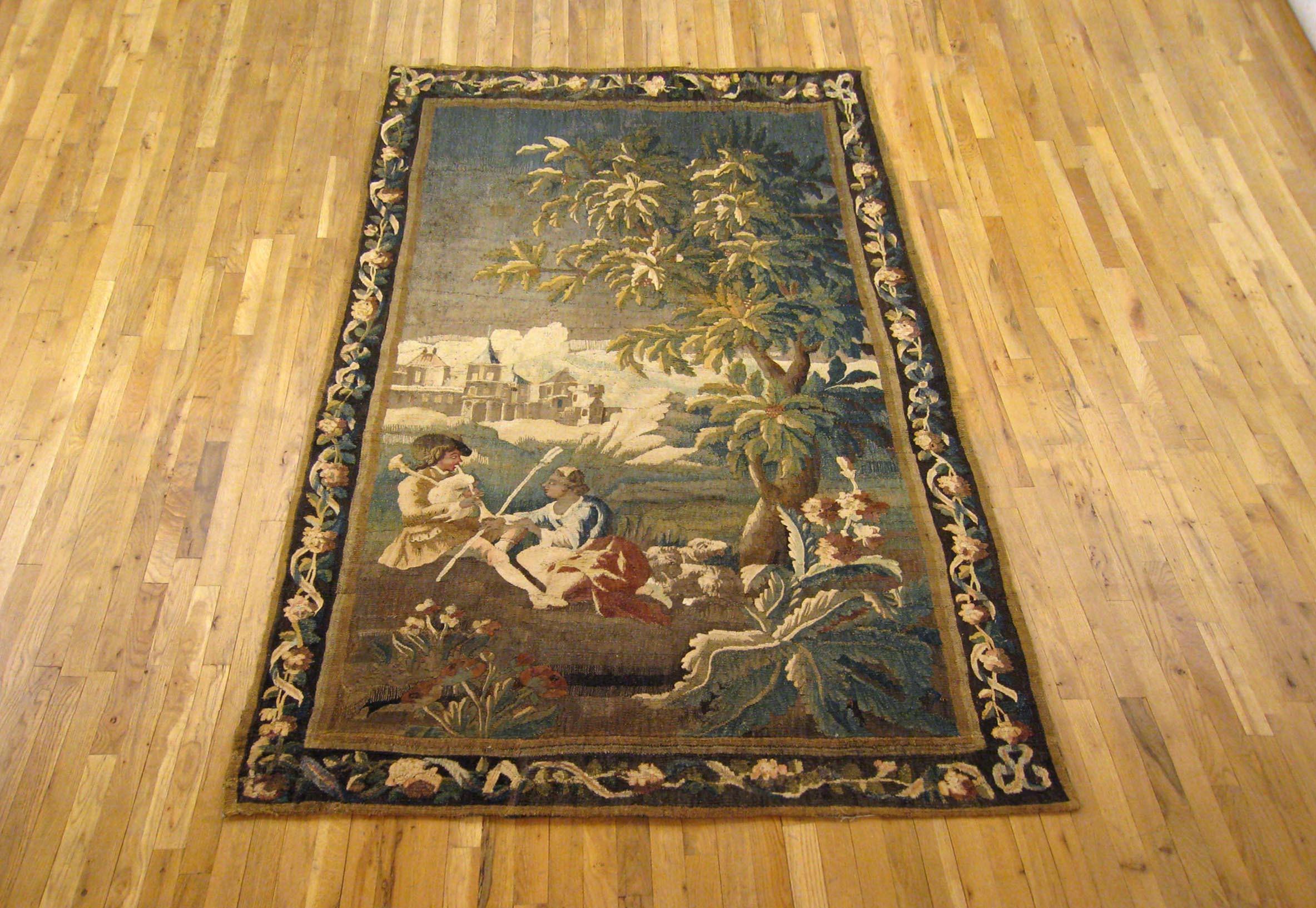 A French Felletin pastoral tapestry panel from the first half of the 18th century, depicting a seated youth playing the bagpipes and courting a seated young woman, flanked by two sheep, within an extensive landscape with a tree to the right-hand