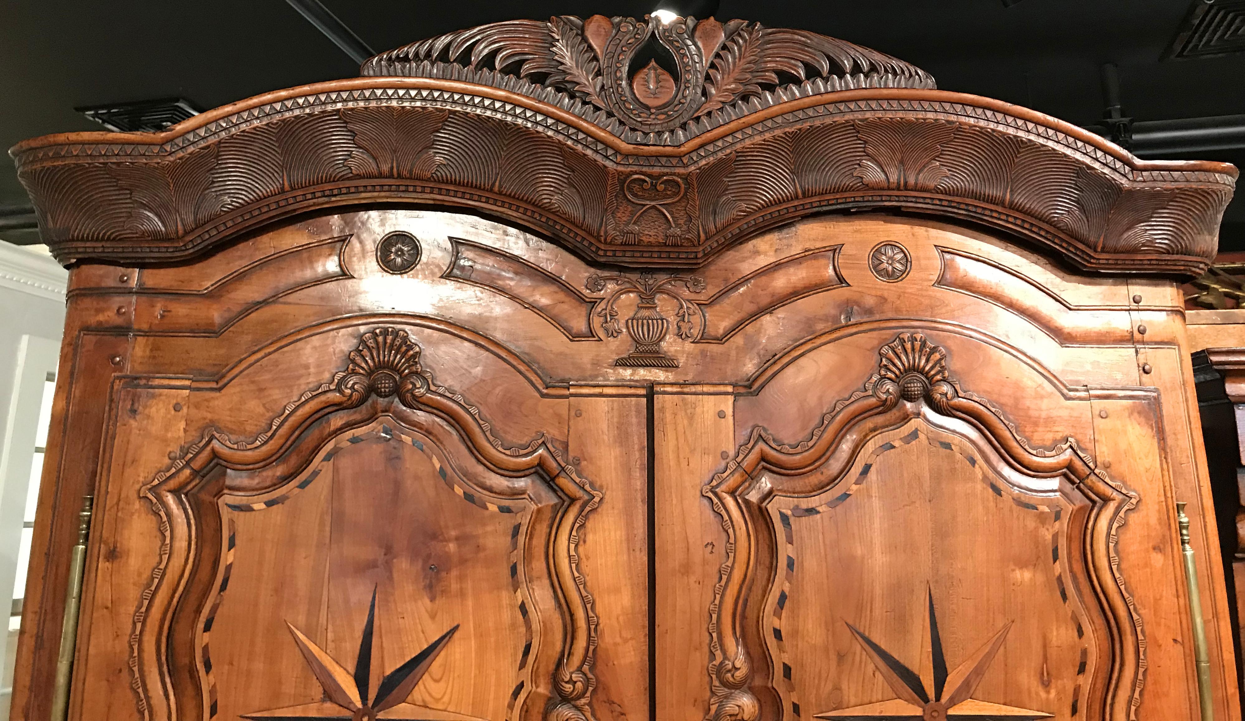 A beautiful French fruitwood armoire with removable double arched fern carved crest surmounting two arched doors, with ornate brass escutcheons and hinges, each exquisitely carved with a small crest fan and foliate decorated scrolled panels - each