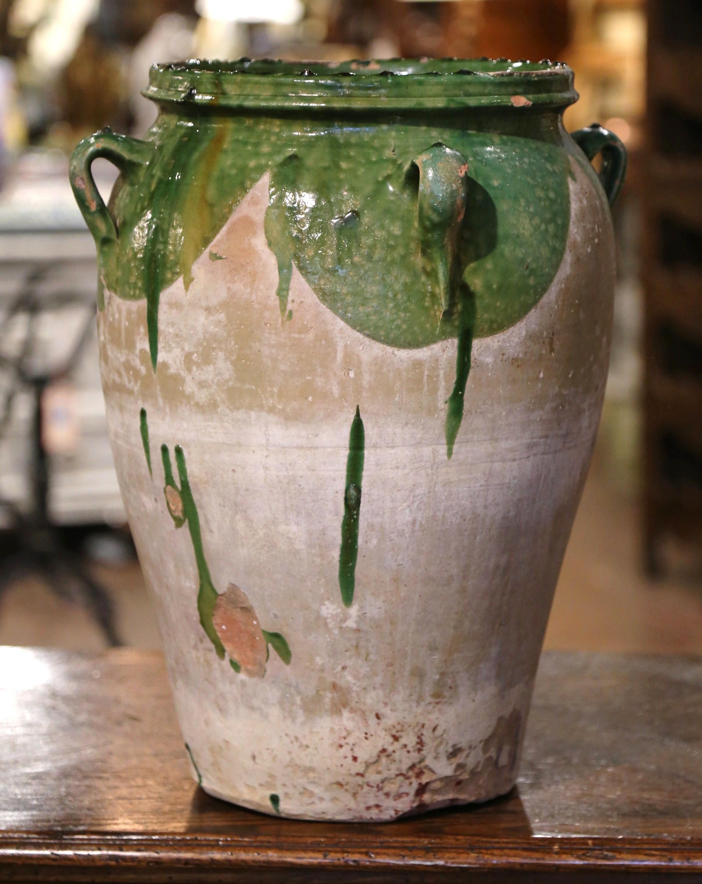 Cette ancienne jarre à olives en faïence a été créée dans le sud de la France, vers 1780. Fabriqué en argile blonde et de couleur neutre, le vase en terre cuite a une forme traditionnelle ronde et bulbeuse ; il est en outre habillé de quatre anses à