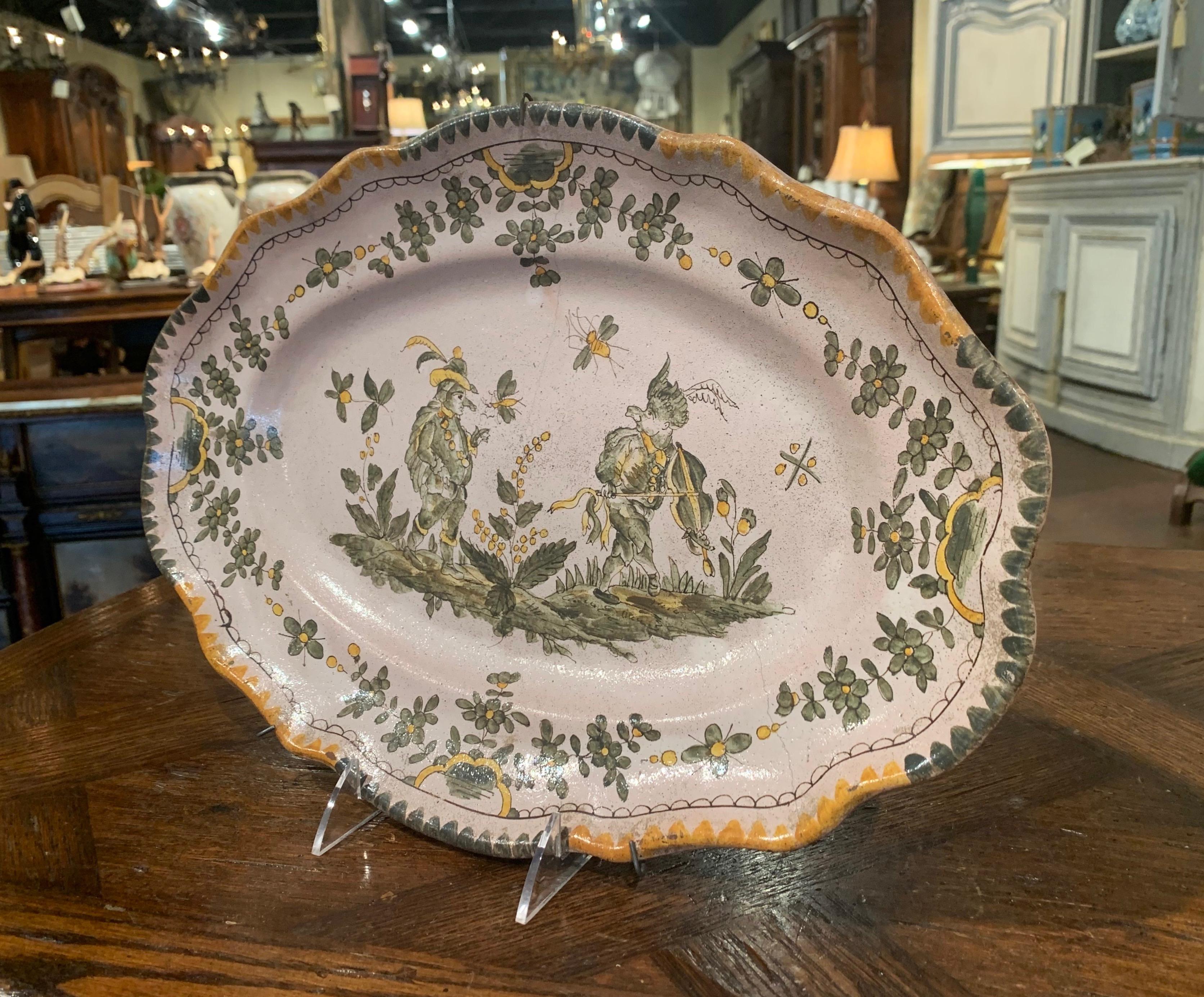 Decorate a wall or a shelf with this colorful antique platter from Provence; crafted in Moustiers, France circa 1780, the oval plate features grotesque people and is hand painted in the soft green and yellow palette. The pottery platter is in