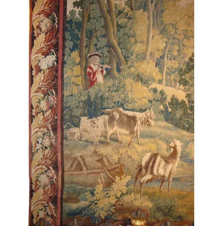 This large and elegant antique tapestry was crafted in Aubusson, France, circa 1760. The colorful wall piece features a country garden scene with goats, architectural elements and a flute player on the left side, all surrounded by foliage, trees and