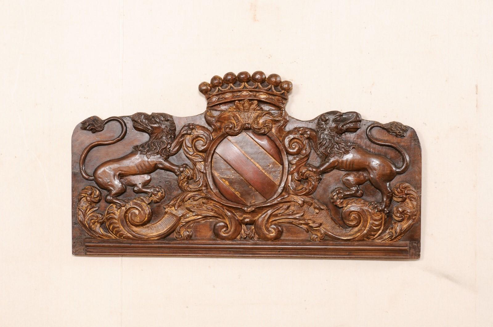 A French carved-wood panel from the 18th century. This antique wall decoration from France has a primarily rectangular shape, with raised carvings depicting a raised crown crest at top center, above a shielded crest at middle, flanked within a pair