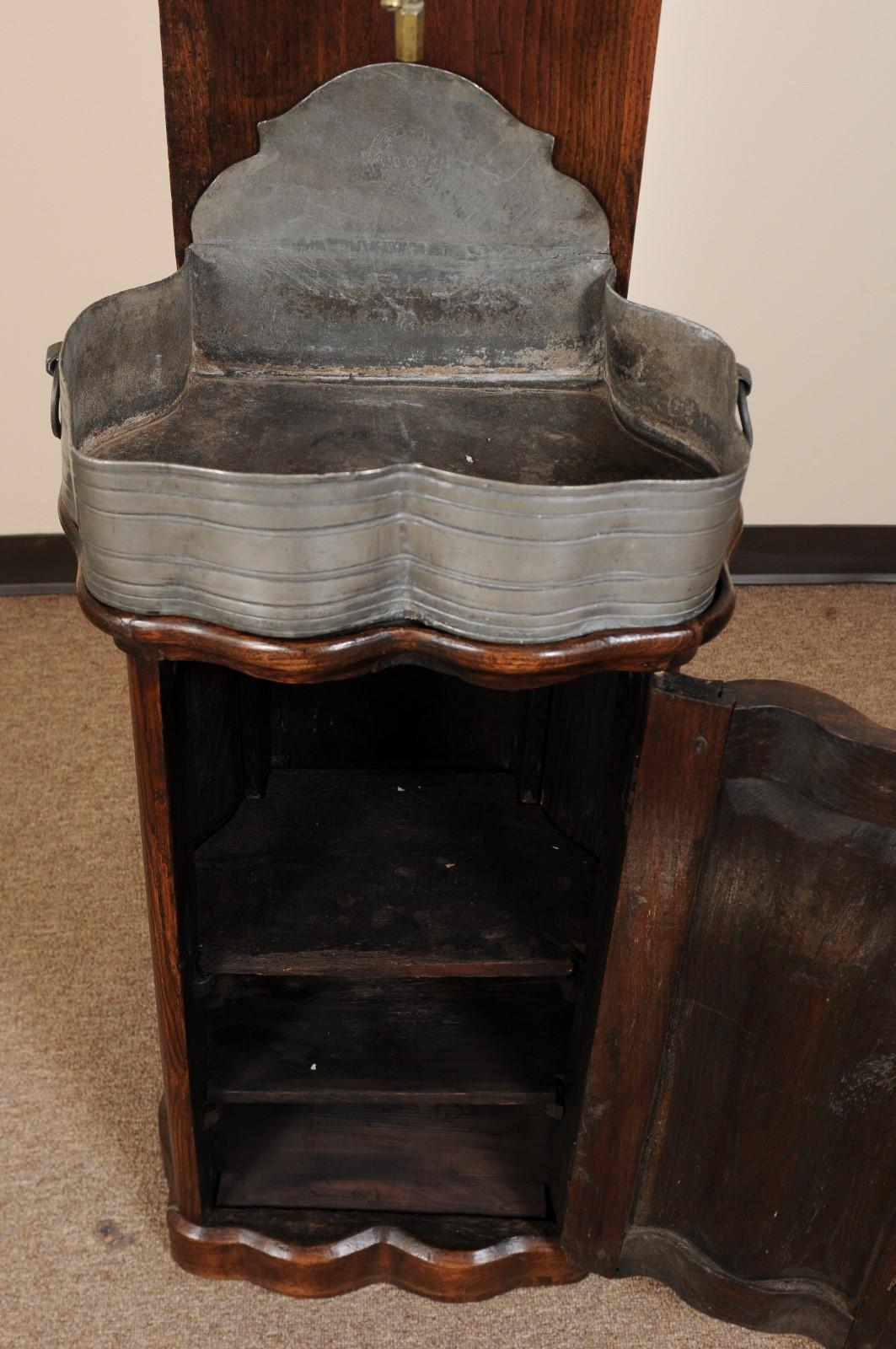 A French 18th century Lavabo in oak featuring shell carving above mounted pewter water container and basin with gable shaped cabinet below. 

 

