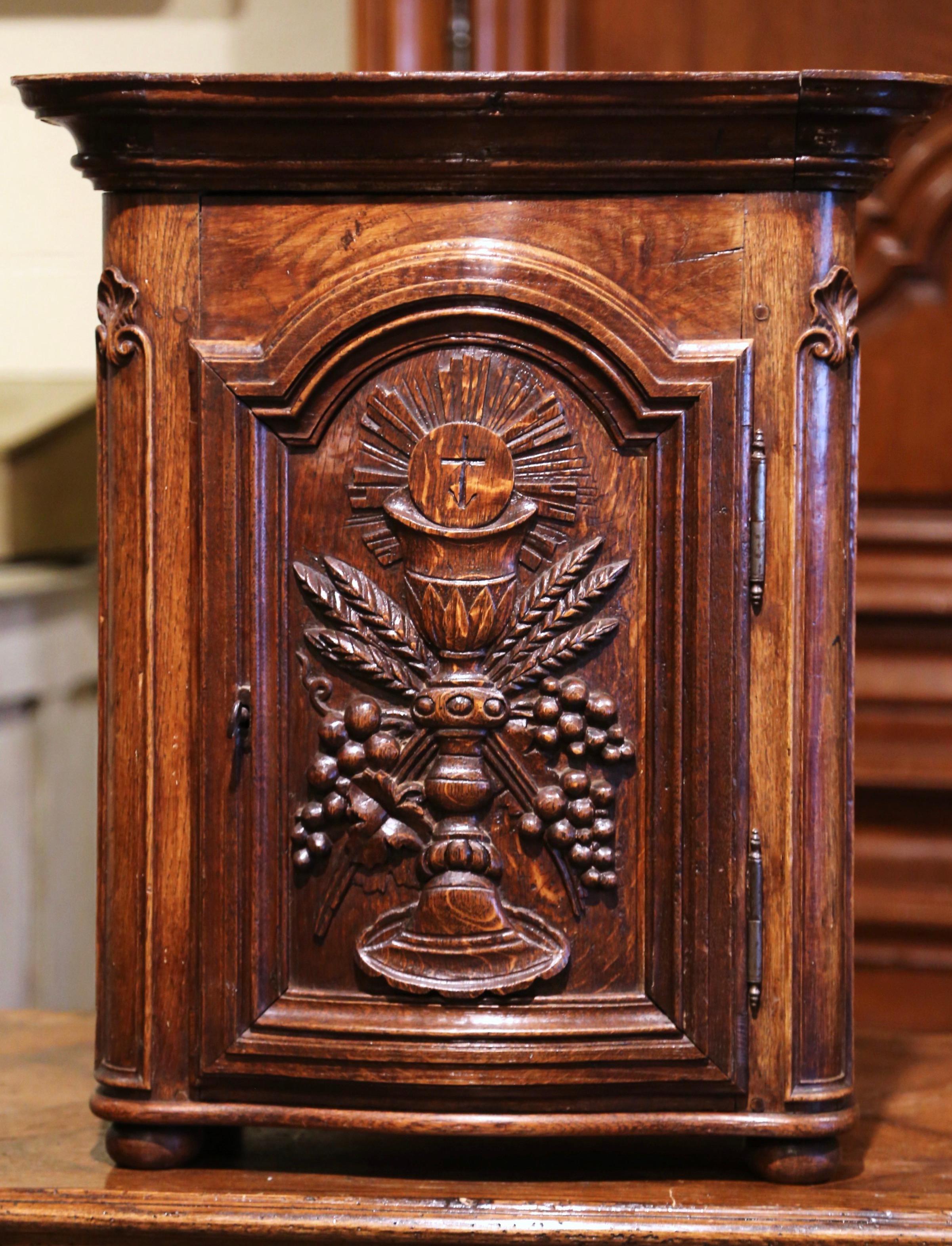This elegant and petite antique church cabinet was crafted in France, circa 1780; the cabinet stands on small bun feet and features a single door with arched top decorated with hand carved catholic motifs including a center monstrance with grape,