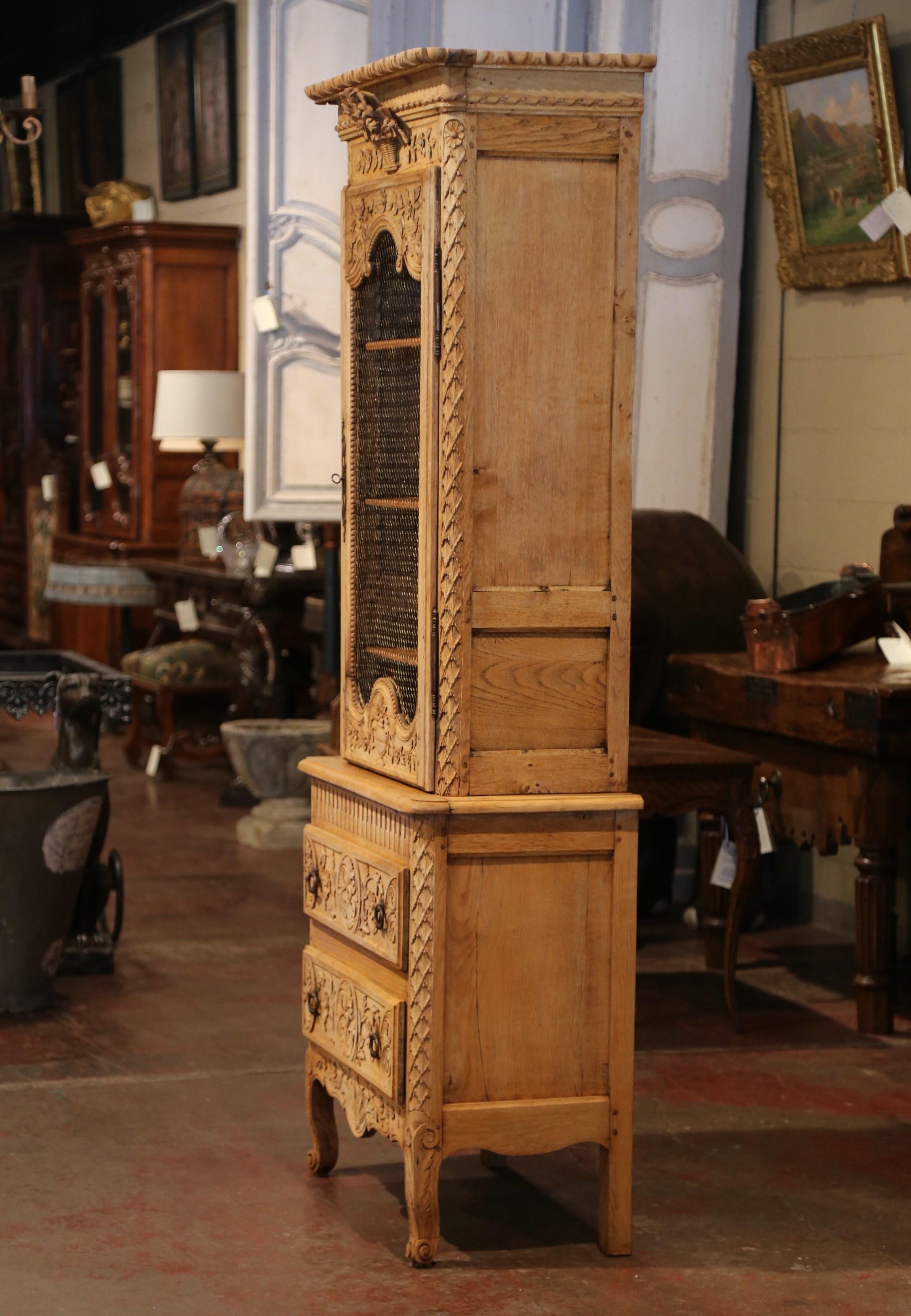 Display a faience or Majolica collection inside this highly hand carved display cabinet. Crafted in Normandy, France, circa 1790, the antique bonnetiere is built in two sections. The top with a beautifully carved floral basket at the pediment,