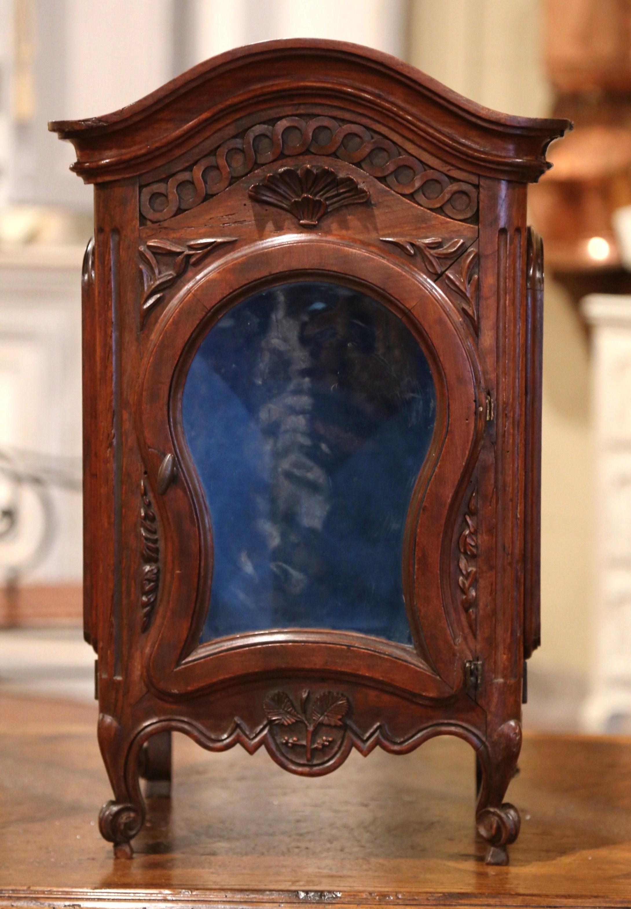 This elegant and petite antique vitrine was crafted in France, circa 1780, the hanging cabinet with bonnet top and shell motif, stands on four escargot feet over a scalloped apron decorated with hand carved flowers and leaves. The wall cabinet