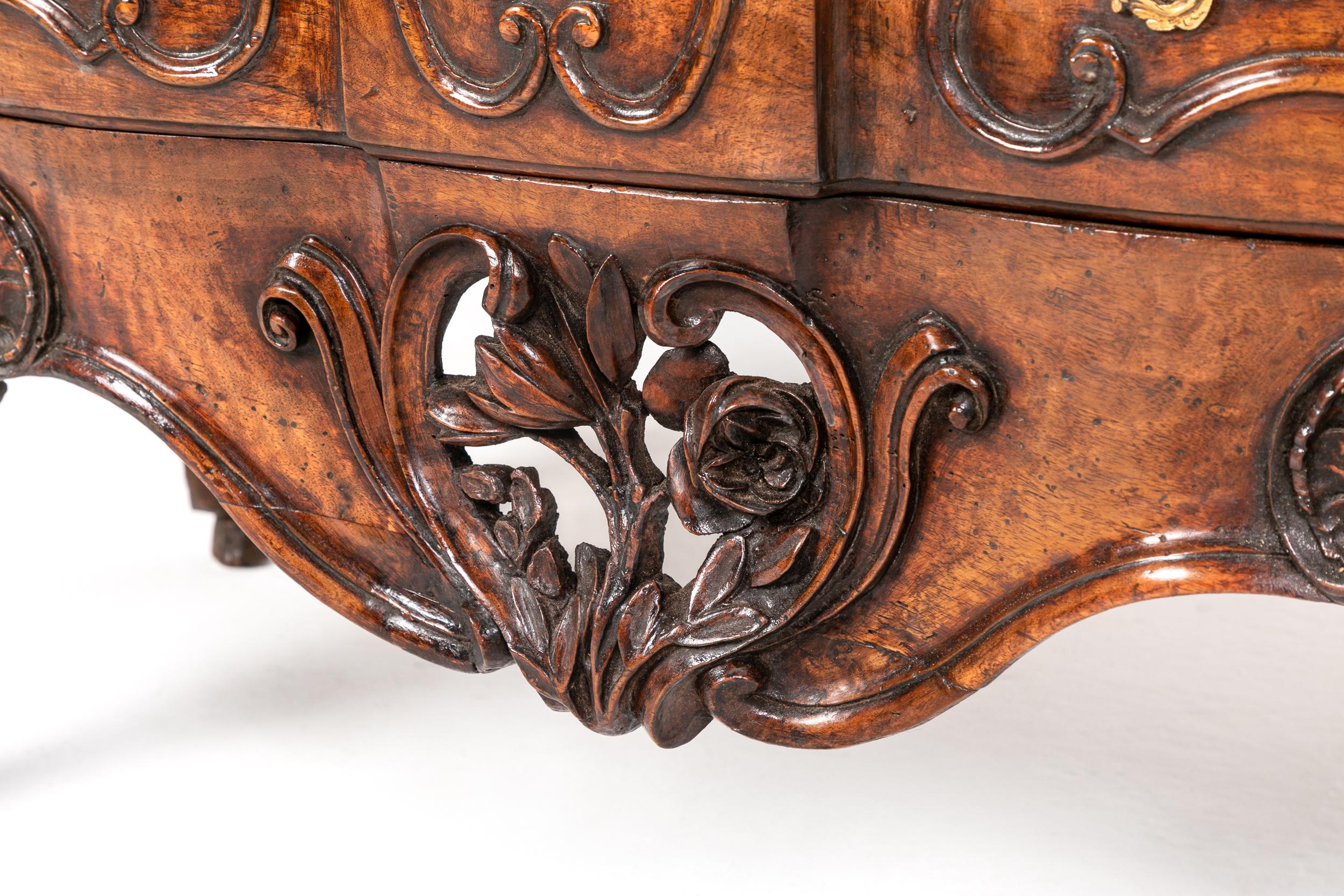 An attractive, 18th century solid walnut commode of the Louis XV period. Having moulded top, and curved sides, with a slight serpentine front. Two drawers with elaborate handles within panels of hand-carved decoration. At the centre is an elaborate