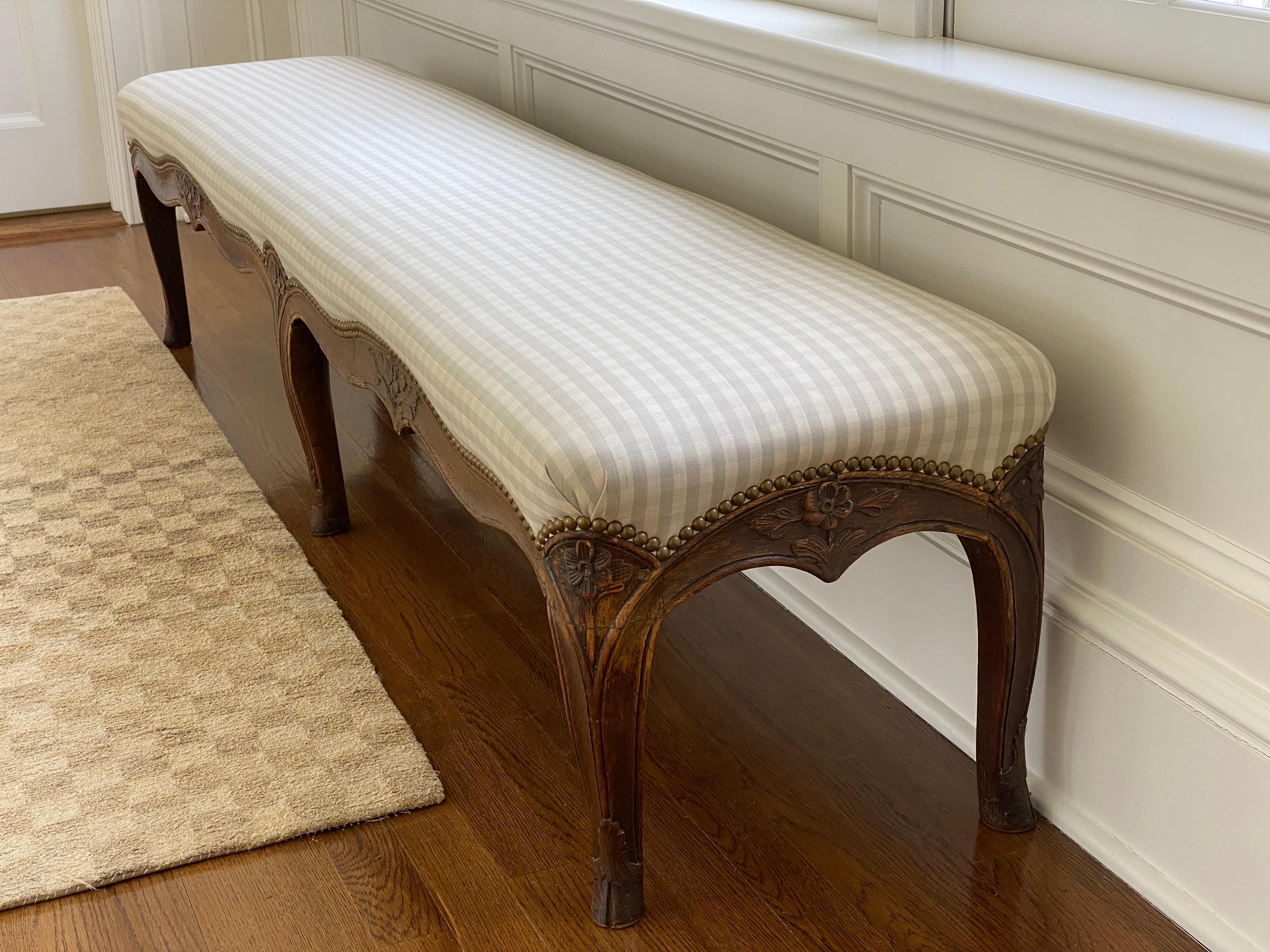 19th century French Louis XV upholstered bench
Hand-carved in Oak, decorated with floral motif on curving cabriole legs. Aged brass nail heads along perimeter. Grey & White check fabric in very good condition. 
Some chips and loss to carving,