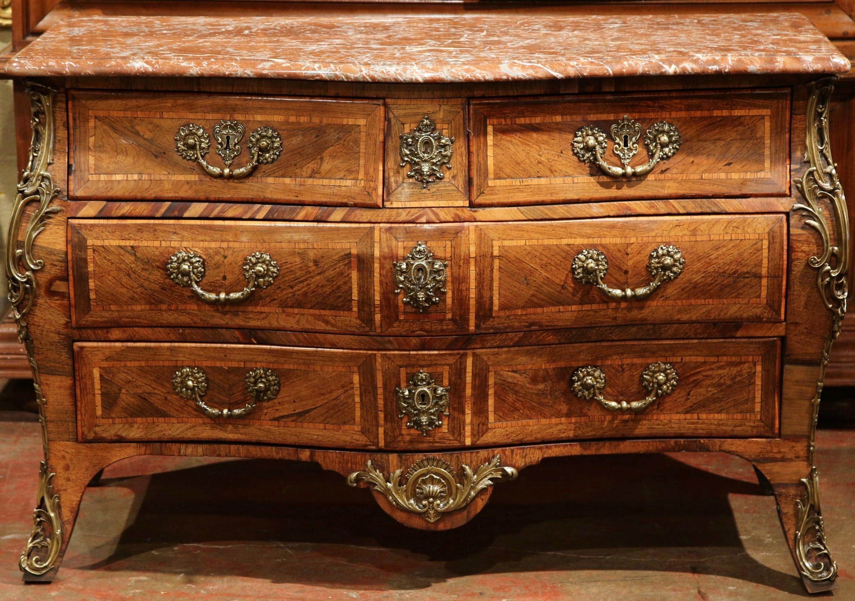Crafted in Burgundy France circa 1750, the antique fruitwood commode stands on curved feet ending with bronze mounts over a he scalloped apron decorated with a bronze shell and leaf motif mount. The chest features three rows across with four bombe