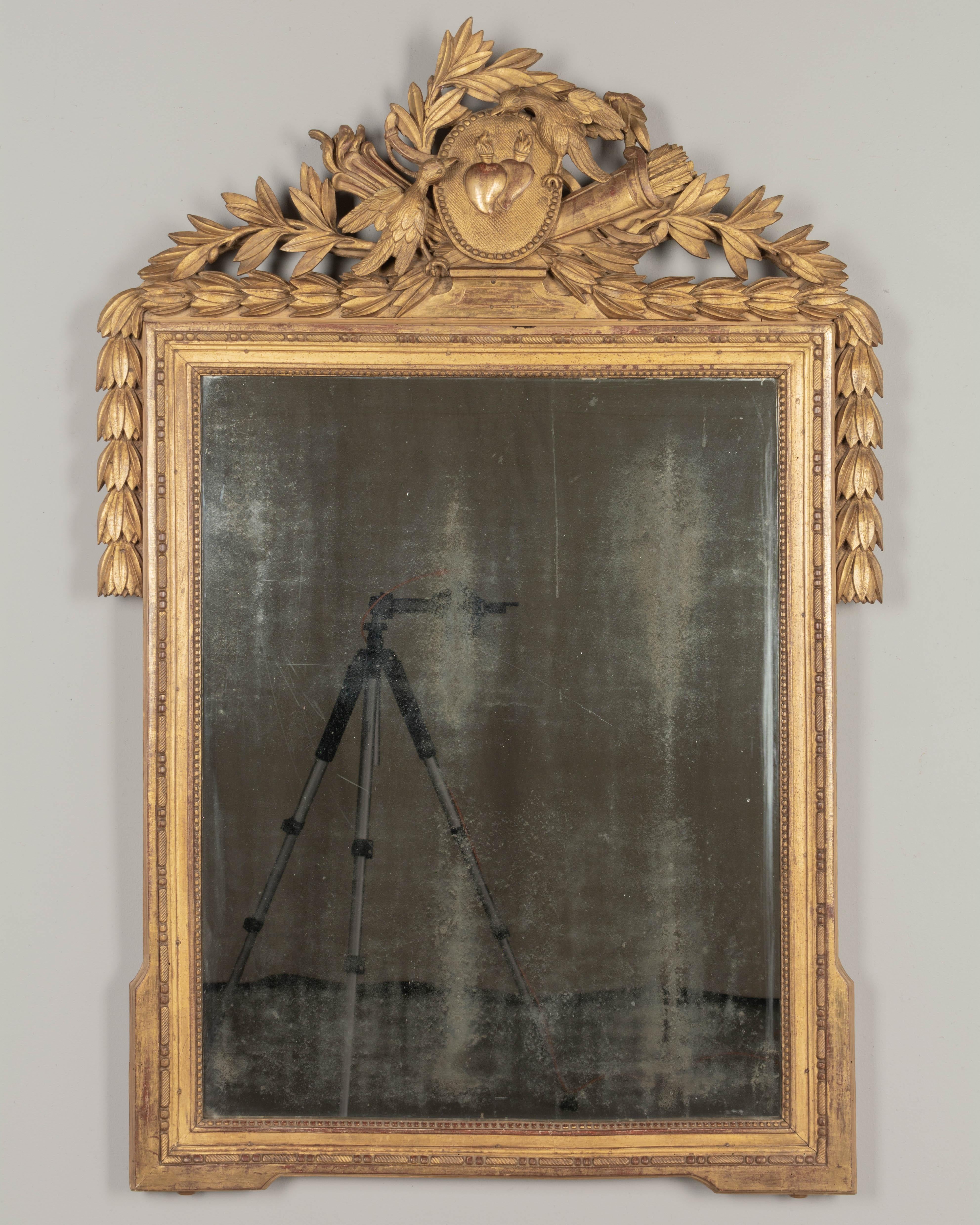 An 18th century Louis XVI French Provençal giltwood bridal mirror. Beautifully carved three dimensional crest with a pair of lovebirds, an oval plaque with flaming hearts and a quiver of arrows. Laurel garlands drape down the sides. Retaining a pale