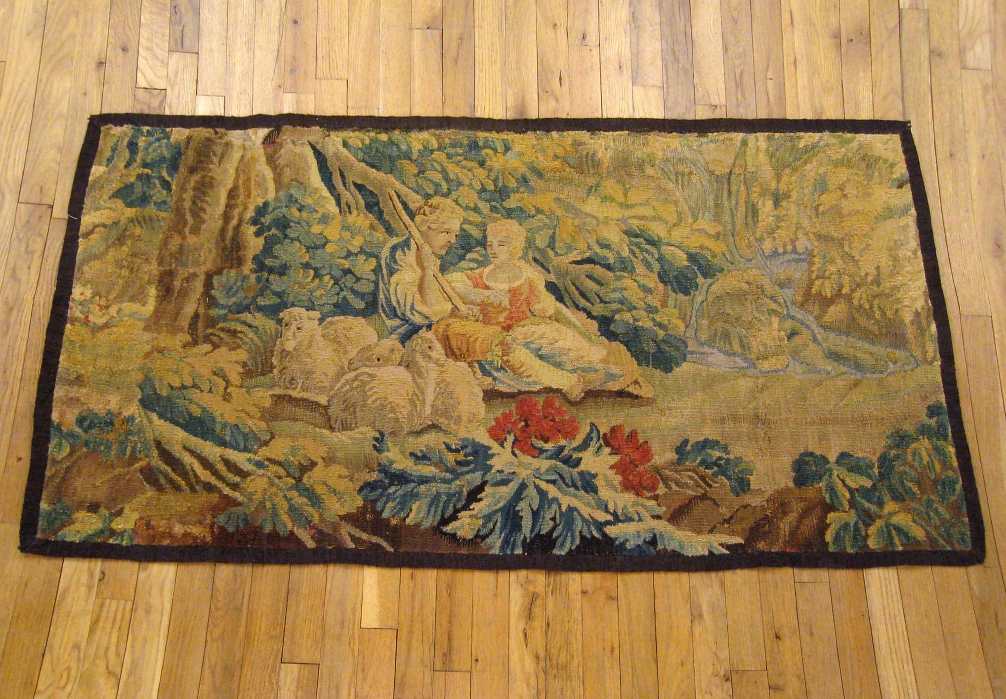 A French pastoral landscape tapestry from the 18th century, envisioning a placid scene in which a shepherd and shepherdess repose under a large tree with three of their sheep within an idyllic forest setting, with a waterfall and stream in the right