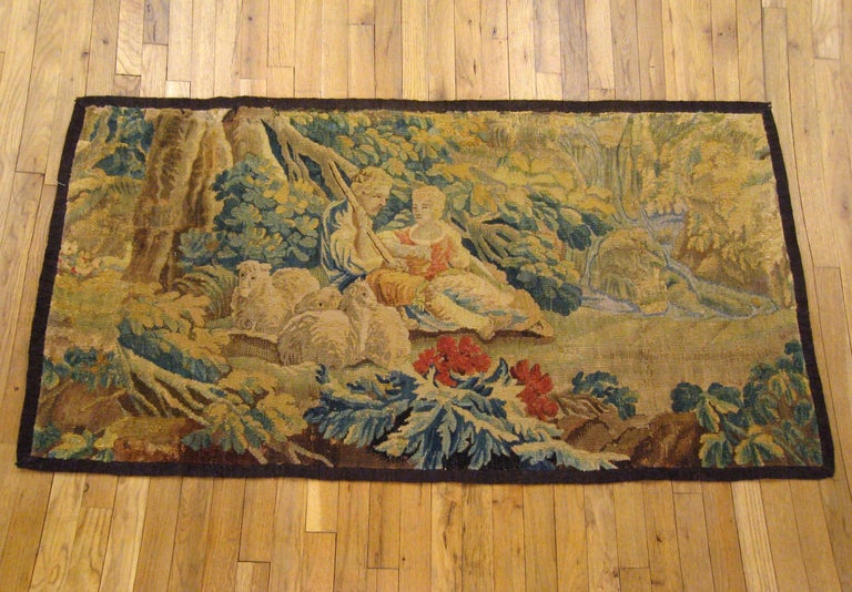 A French pastoral landscape tapestry from the 18th century, envisioning a placid scene in which a shepherd and shepherdess repose under a large tree with three of their sheep within an idyllic forest setting, with a waterfall and stream in the right