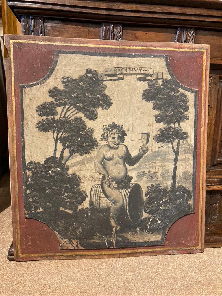 An amusing French Provincial painted panel showing a chubby Bacchus, the Greek god of wine, astride a wine barrel, naked except for grapes and wine leaves around his waist, holding a goblet aloft. The raised cartouche painted in grisaille technique,