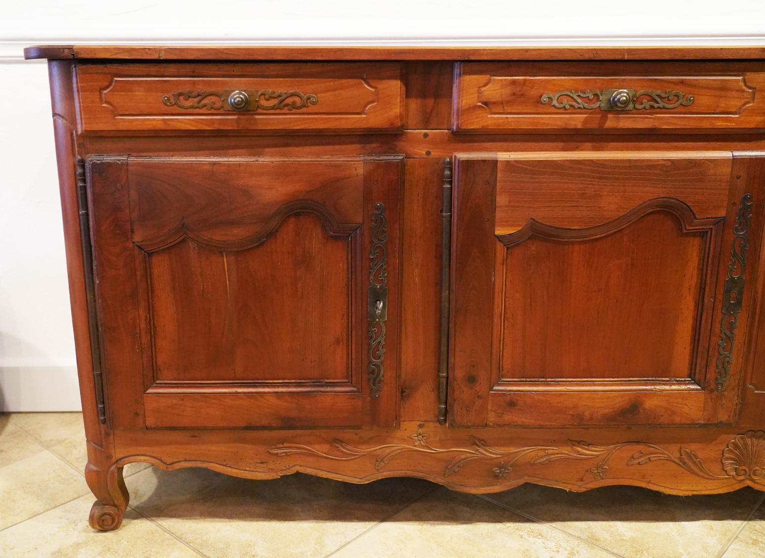 104 inches long this French Provincial carved cherrywood buffet or enfilade features four carved panel doors and four drawers above. It rests on four carved feet connected by a beautifully carved and scalloped apron. Hinges, handles and locks are