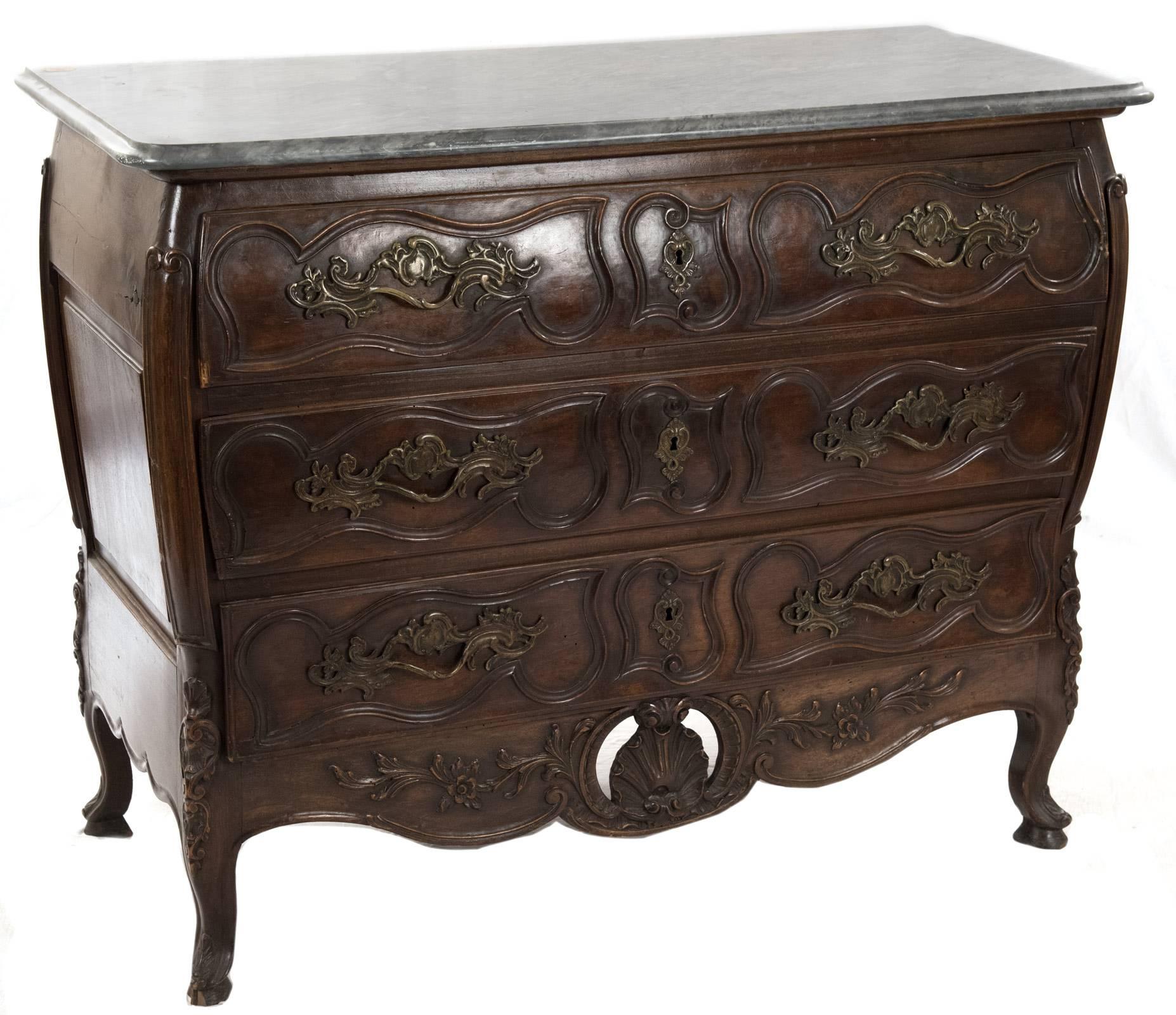 A finely carved late eighteenth century walnut commode in the French provincial Louis XV style particular to Nimes. A rectangular grey marble top sits above a commode of bombe form with carved canted corners flanking three long drawers, each