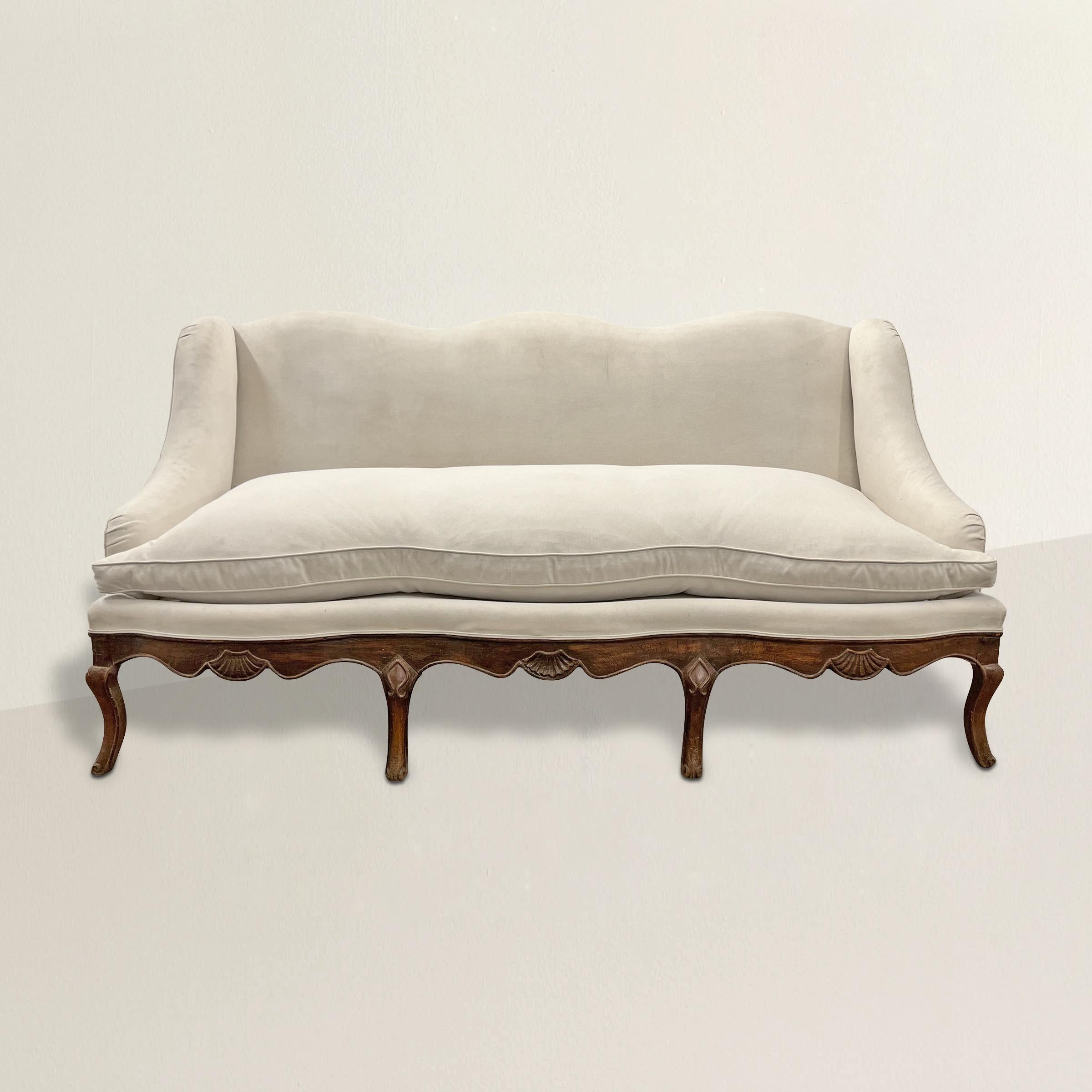 A timeless, curvy, and sophisticated 18th century French Provincial Louis XV walnut framed sofa with a gently reclined back with a scalloped top, a scalloped down-filled cushion, sloping arms, carved shell cartouches on the apron, and all resting on