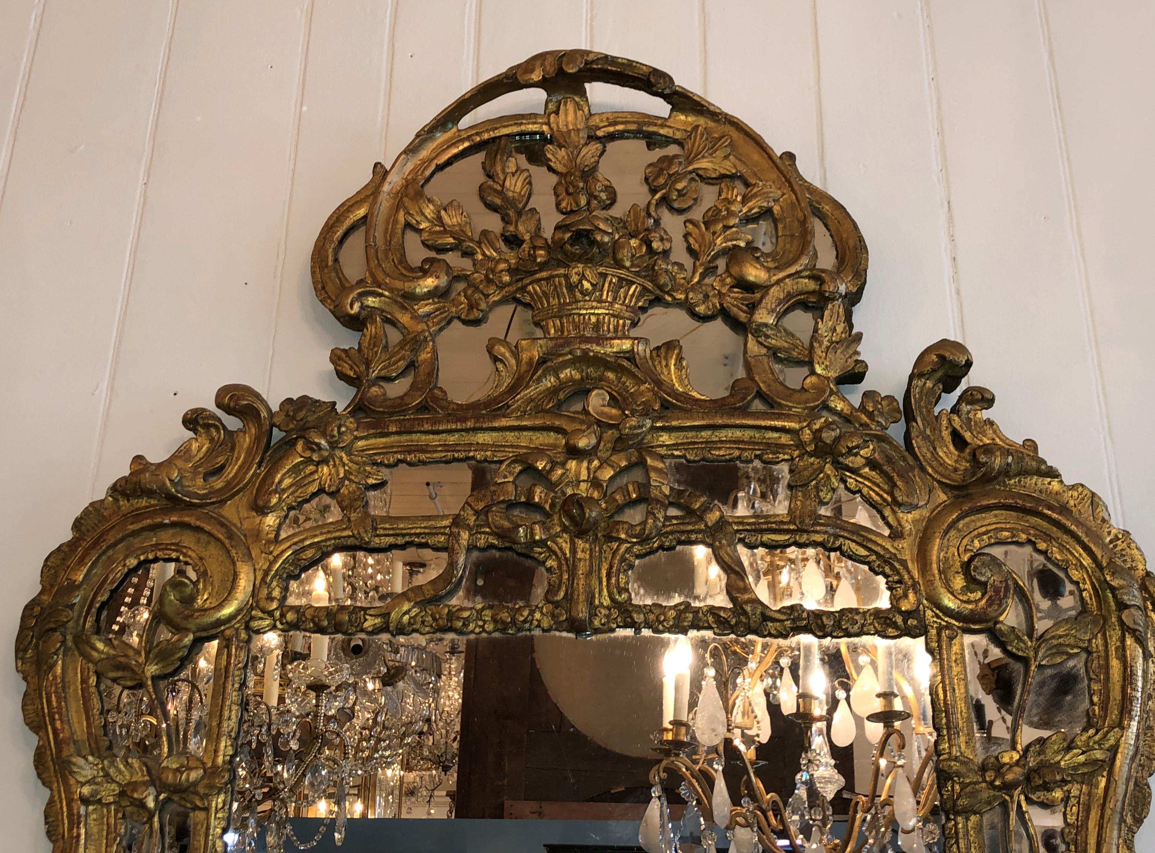 This elegant 18th century Régence looking glass has carved giltwood with mirrored borders, circa 1715-1730. The carved frame has floral and scrolling elements laid over the mirrored borders and crest. The pediment has a carved basket framed by