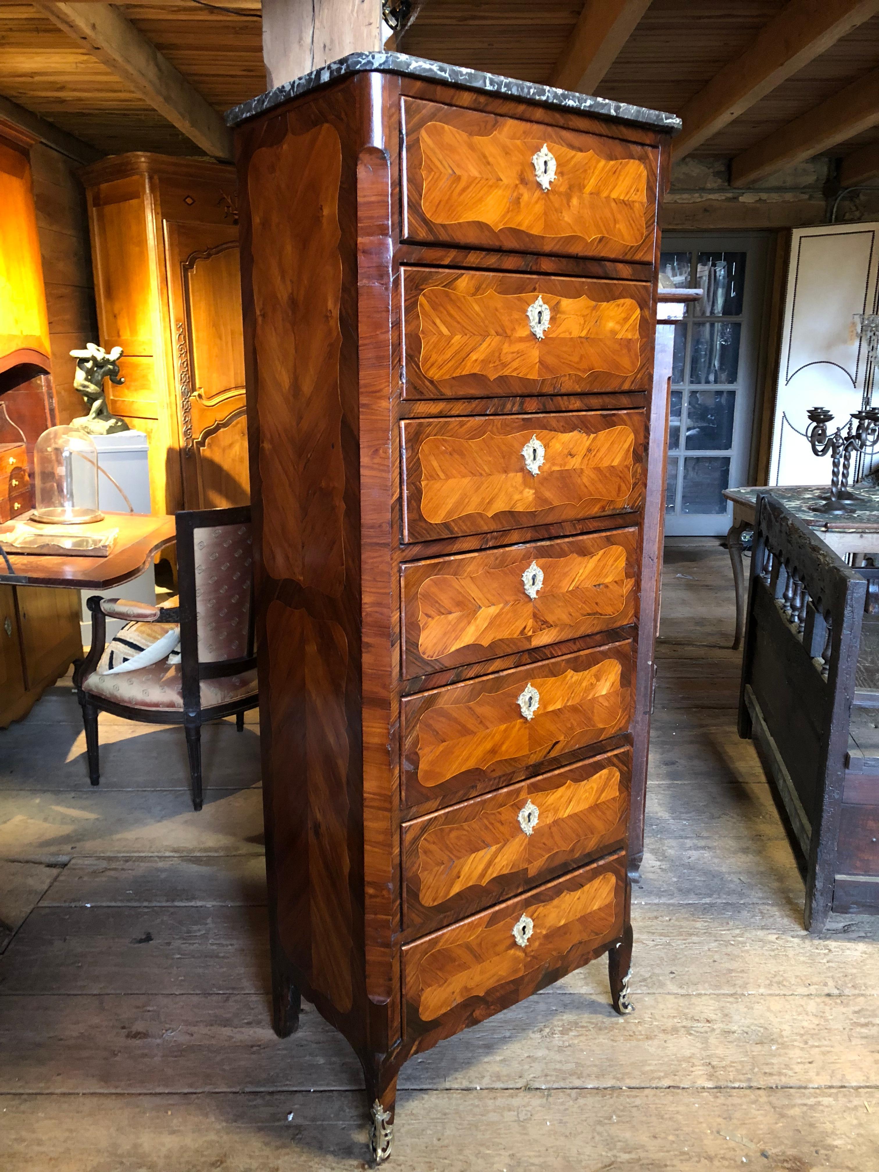 A French Louis XV period semainier (7-drawer chest) signed on top front left under stone top by Jean-Baptiste Tuart (maitre 1 Feb. 1741) in hardwood marquetry veneer over oak and other secondary woods, retaining its original grey marble top and gilt