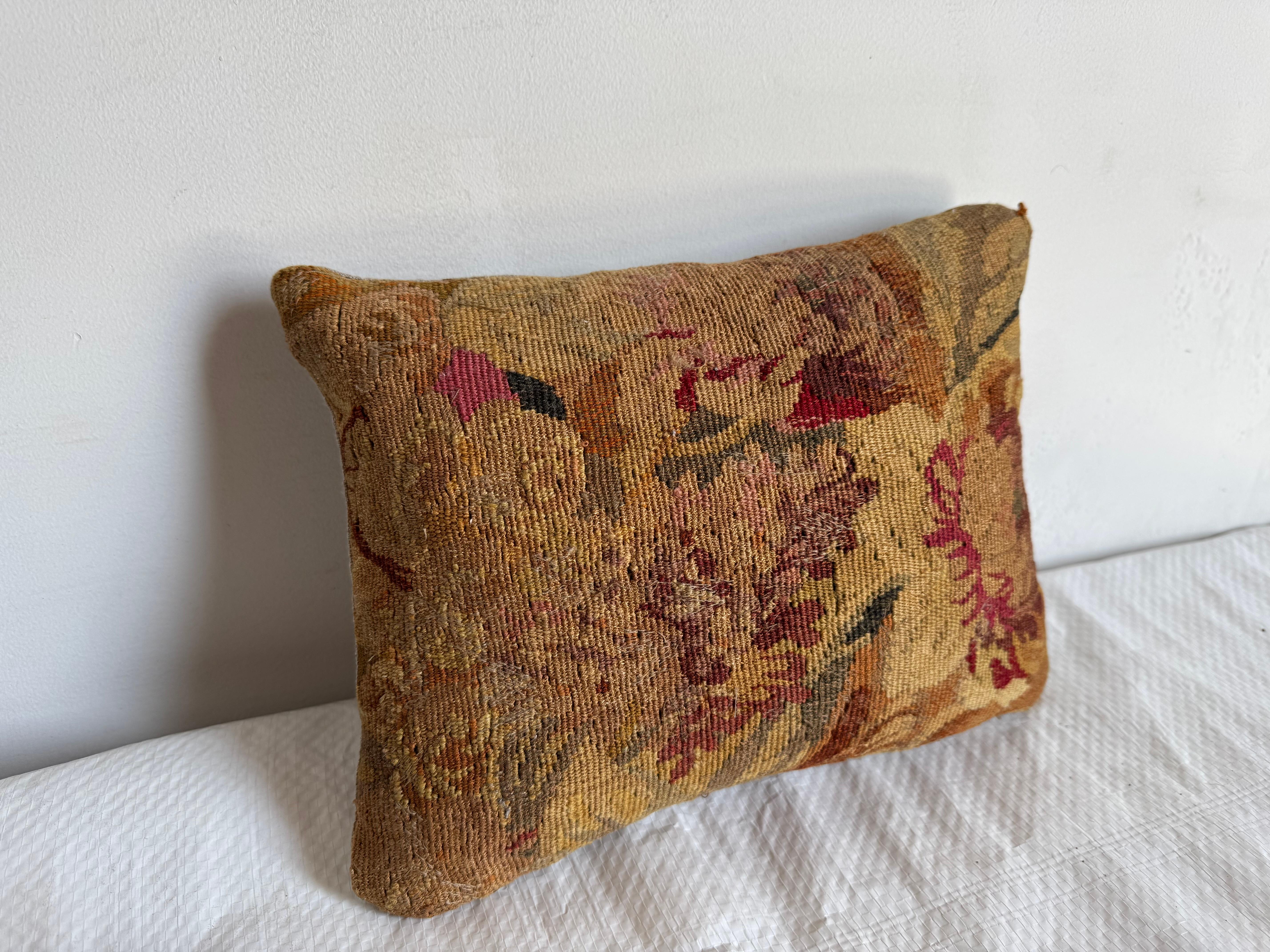 Se trata de un cojín tapiz vintage de estilo provincial francés con una estética desgastada y rica en texturas. El cojín presenta un intrincado motivo floral en una paleta de dorados apagados, rojos intensos y verdes oliva, tejido sobre un fondo de